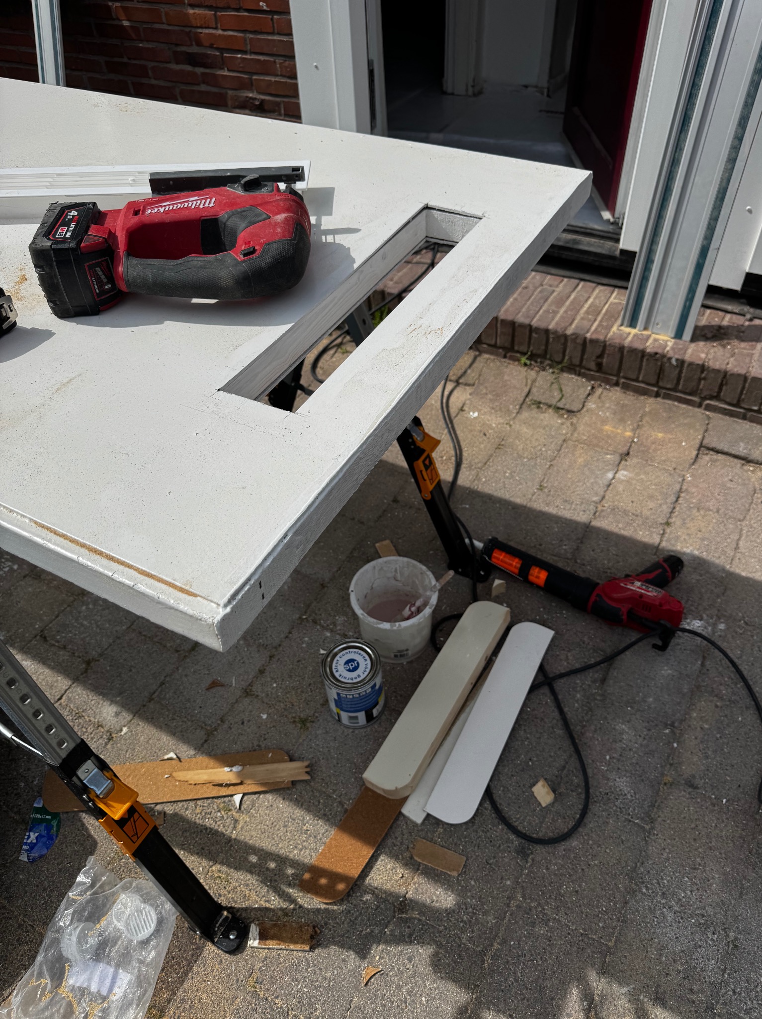 Białe drzwi z wyciętym otworem, piła akumulatorowa Milwaukee, puszka farby i listwy przypodłogowe na zewnątrz, prawdopodobnie podczas montażu lub naprawy.