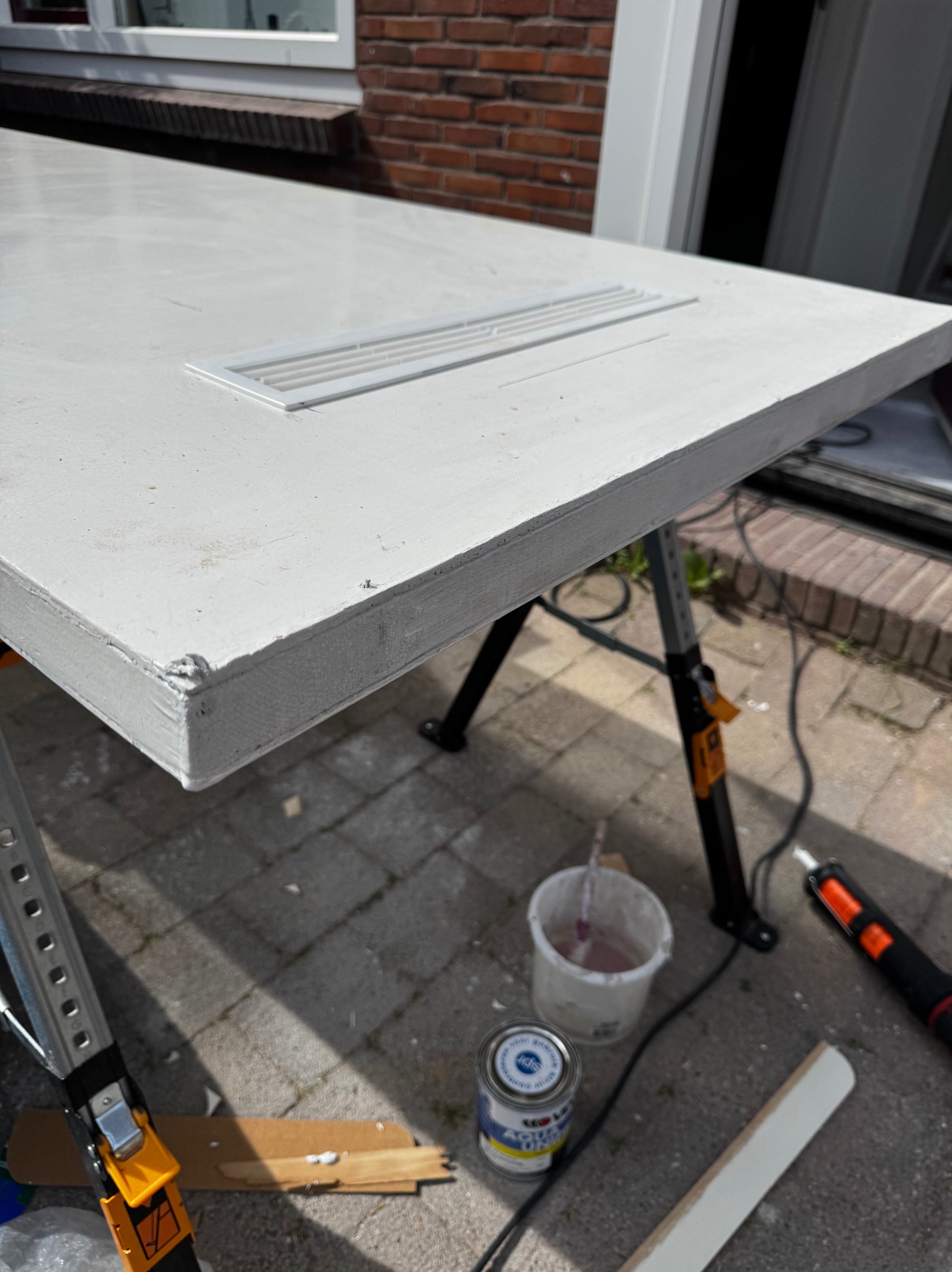 Białe, świeżo pomalowane drzwi z kratką wentylacyjną, ustawione na kozłach. Widoczne narzędzia malarskie i puszka farby. Ceglany budynek w tle.