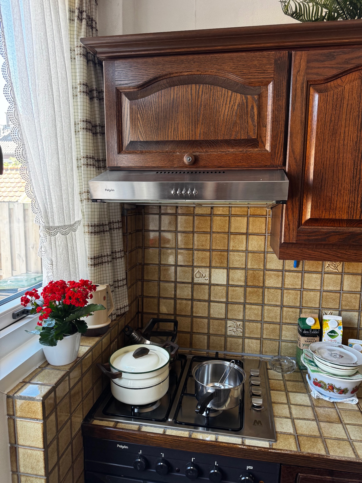 Kuchnia z drewnianymi szafkami, okapem i oknem z firanką. Na kuchence gazowej stoją garnki, a na parapecie kwiaty. Widoczne brązowe płytki na ścianie.