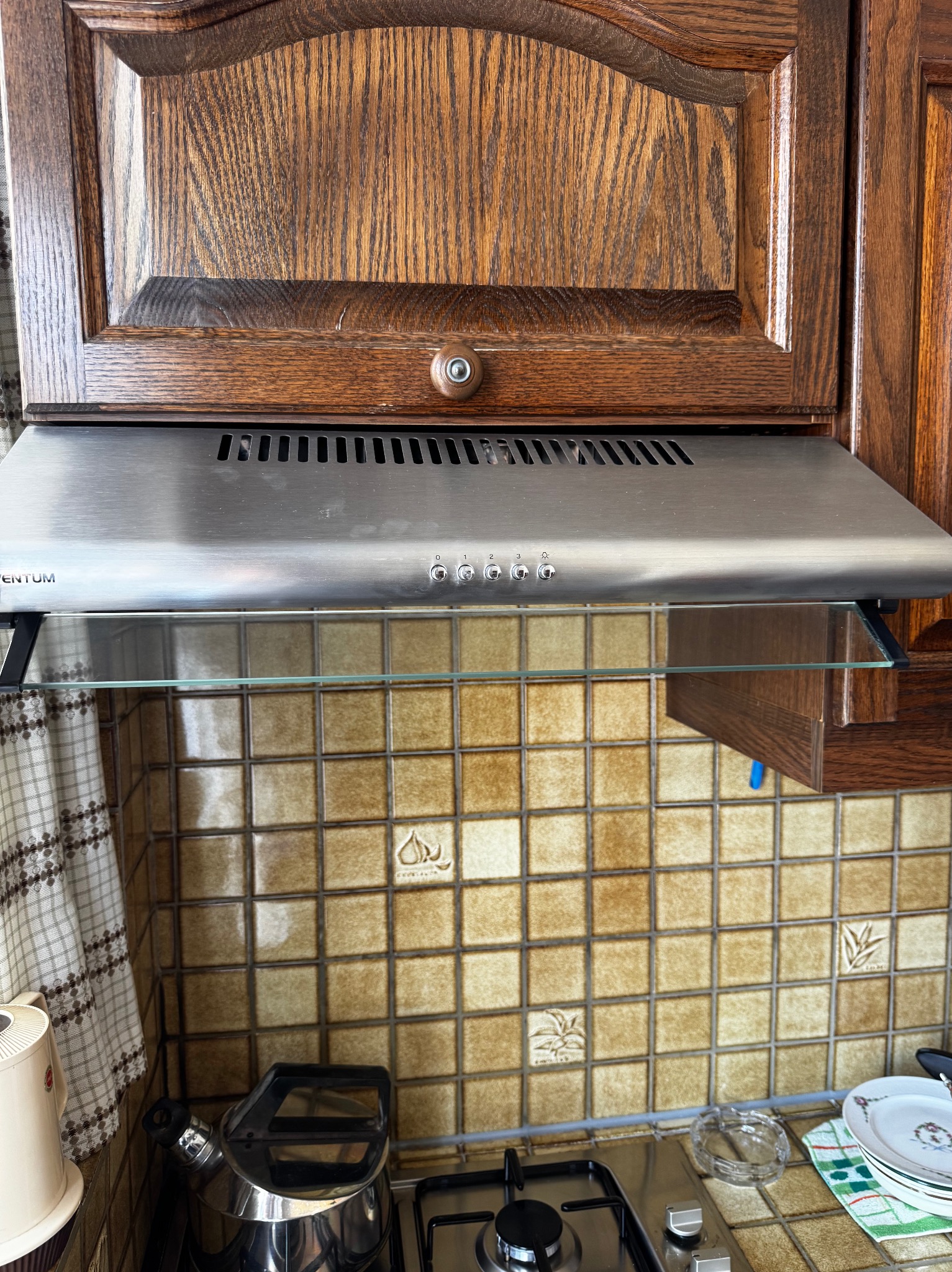 Szczegół ujęcia kuchennego okapu podszafkowego ze stali nierdzewnej, zamontowanego pod drewnianą szafką, z widocznymi płytkami ceramicznymi w tle.