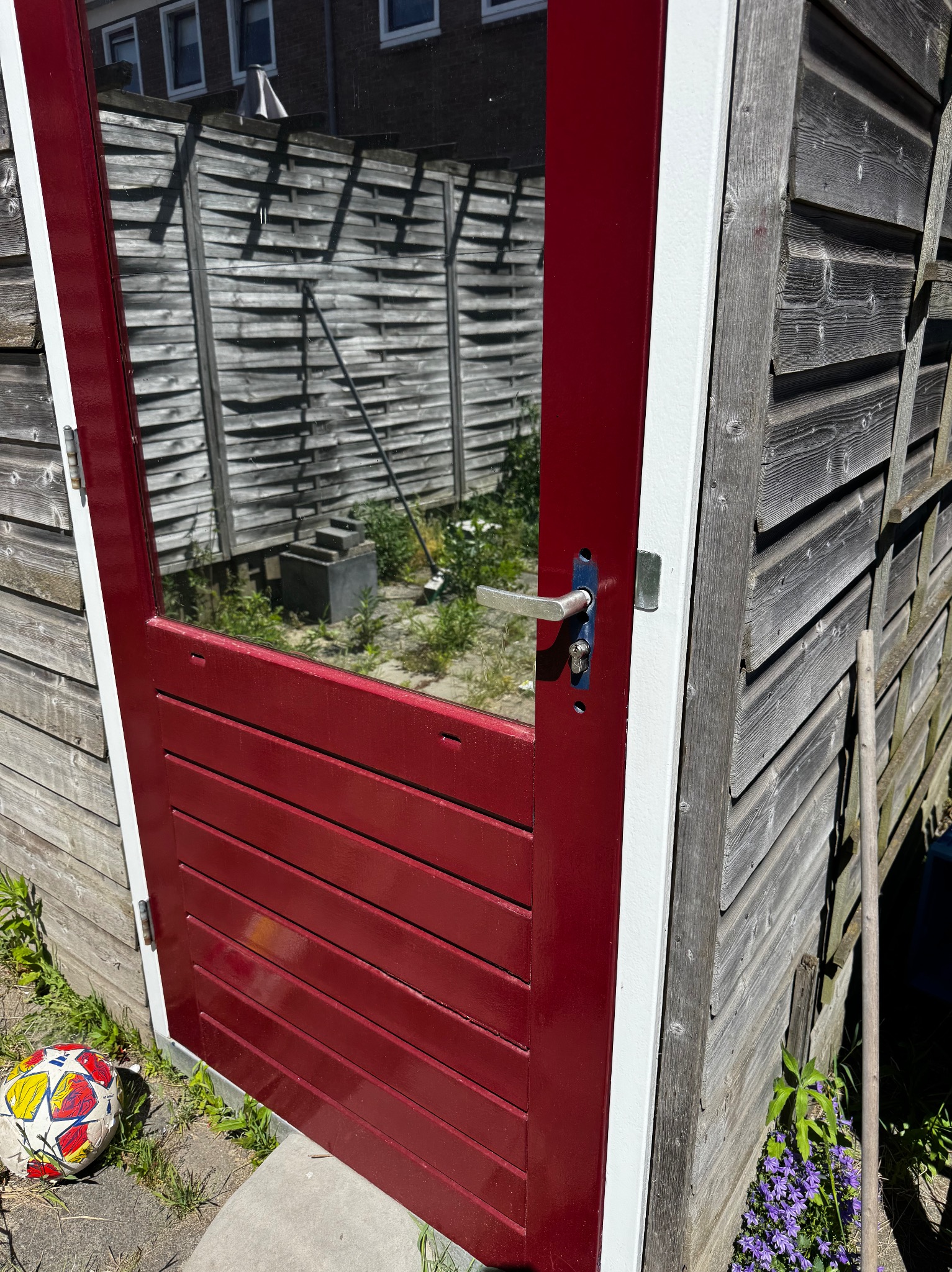 Czerwone drzwi z szybą w drewnianej szopie, widoczne odbicie otoczenia, piłka leżąca u dołu. Nietypowe ujęcie drzwi zewnętrznych.
