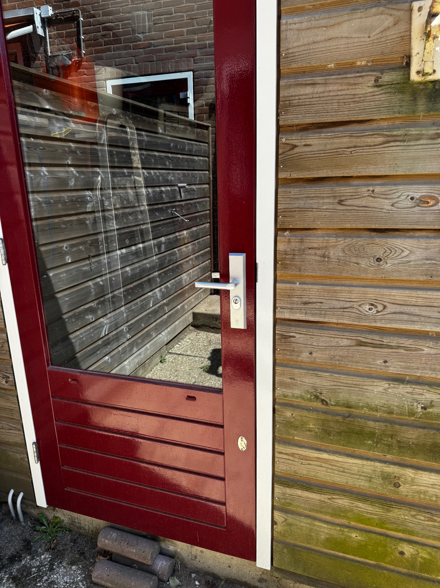 Ciemnoczerwone drzwi z szybą i srebrną klamką, zamontowane w drewnianej ścianie z widocznymi śladami wilgoci. Widoczne cegły pod drzwiami.