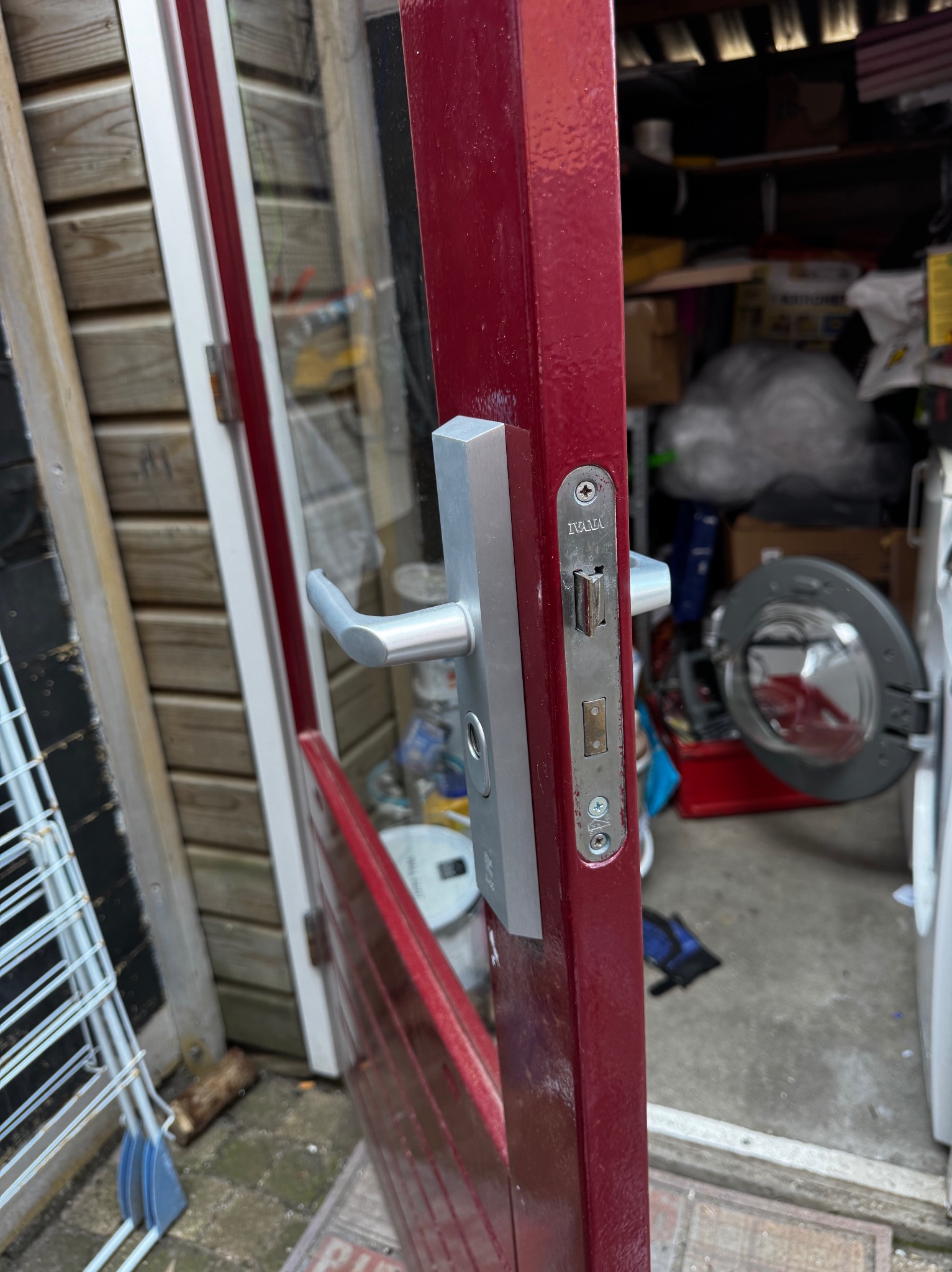 Częściowo otwarte, bordowe drzwi z srebrną klamką i zamkiem IVANA, prowadzące do zagraconego pomieszczenia z pralką. Widoczne ślady użytkowania na zamku.