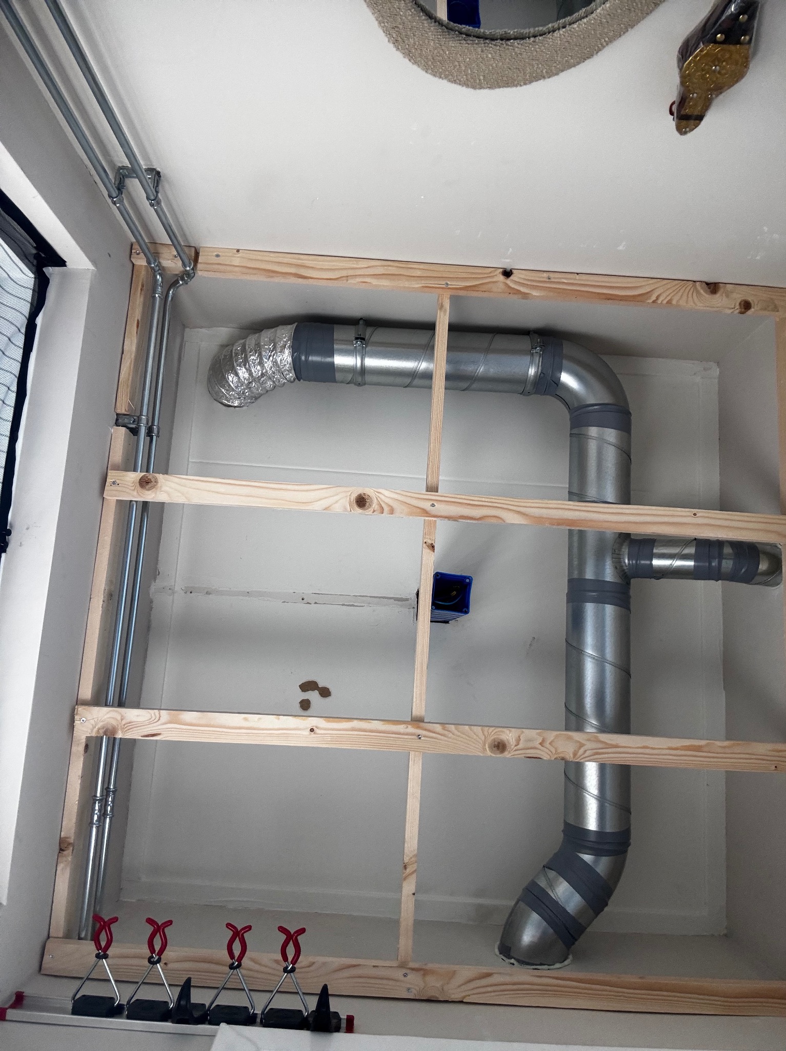 Widok instalacji wentylacyjnej z rurami aluminiowymi w drewnianej ramie, z widocznymi elementami mocującymi i fragmentem okna po lewej stronie.