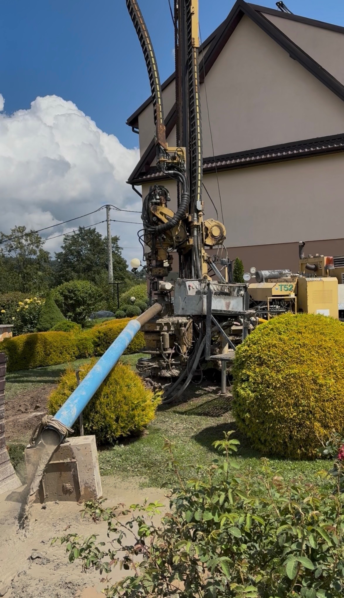 Żółta wiertnica do studni głębinowych w trakcie pracy na tle domu i krzewów. Woda wypływa niebieską rurą. Widoczne detale maszyny i otoczenia.