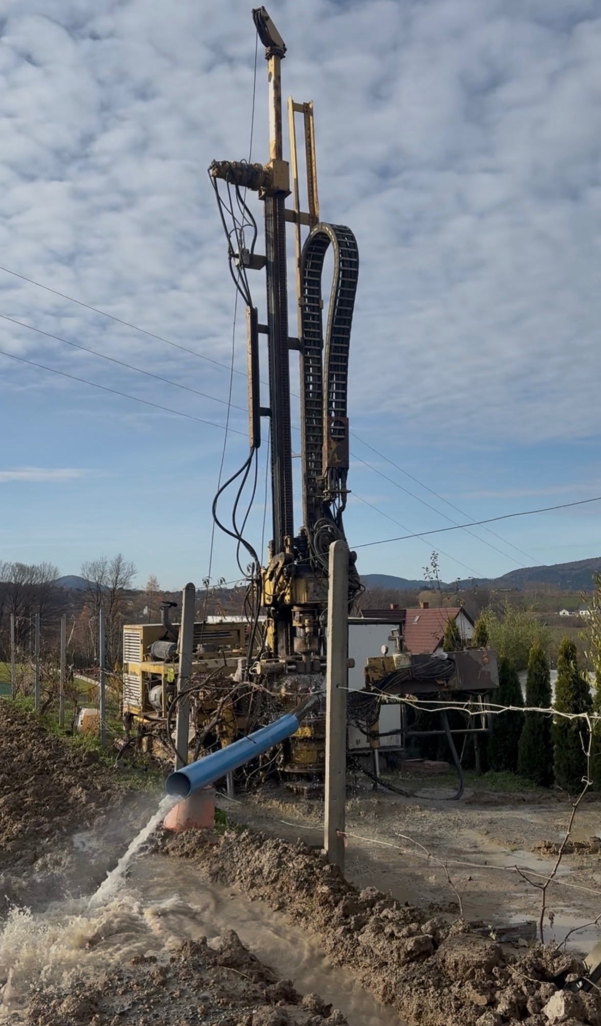 Żółta wiertnica w trakcie pracy; woda wypływa z niebieskiej rury, teren błotnisty, w tle domy i drzewa. Wiertnica na tle pochmurnego nieba.