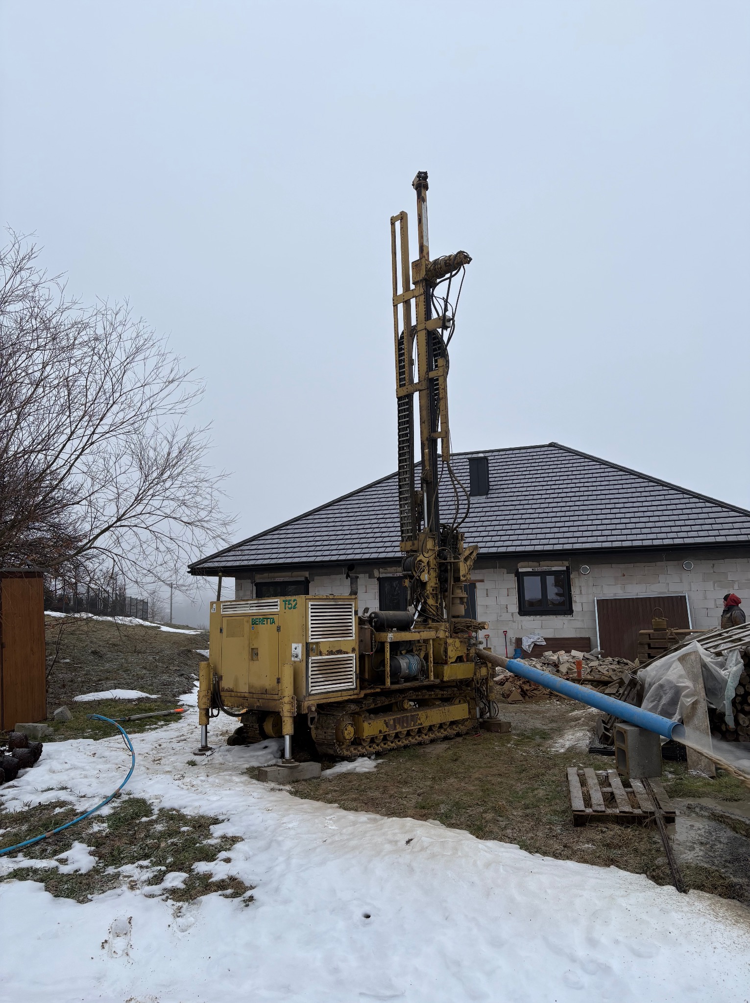 Żółta wiertnica Beretta T52 na gąsienicach, w trakcie pracy przy budynku z szarą dachówką. Wokół resztki śniegu i błoto. Widoczny niebieski wąż i wypływająca woda.
