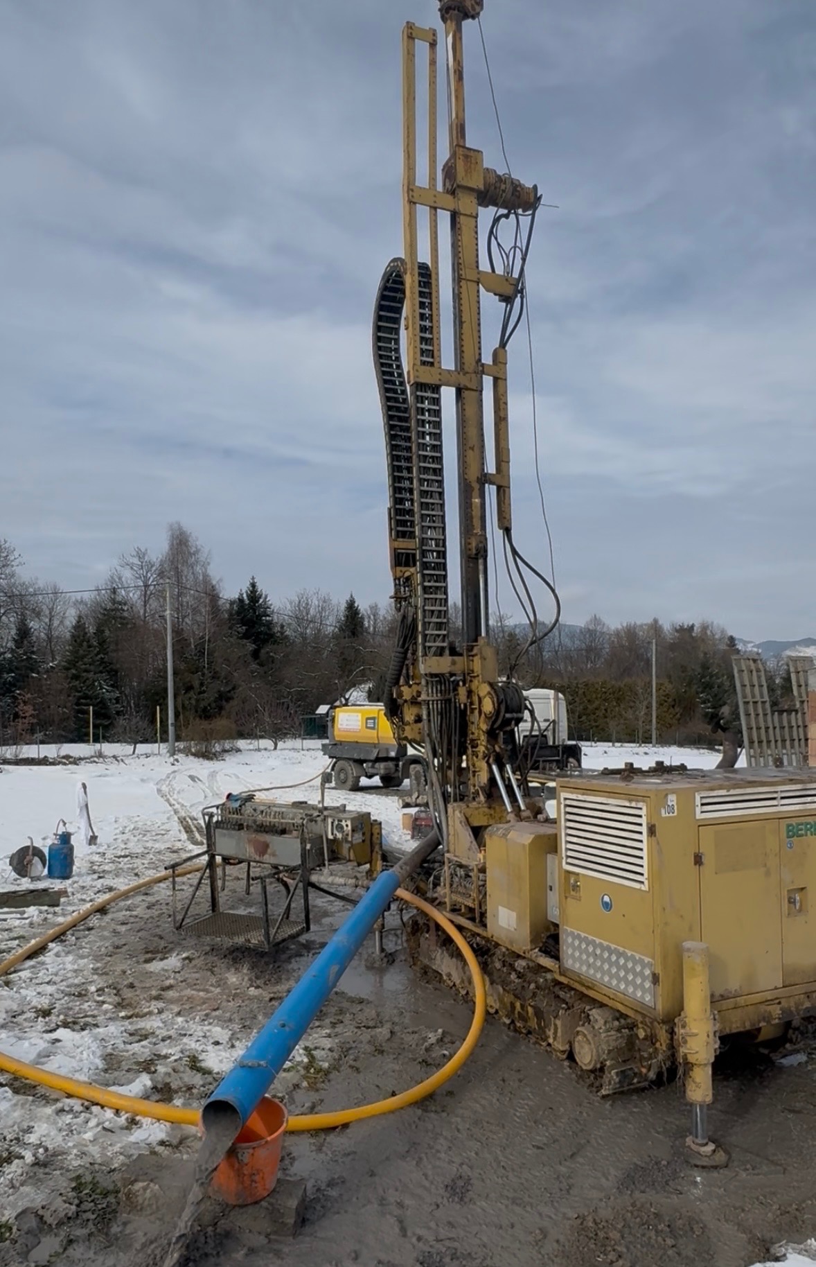 Żółta wiertnica w trakcie pracy na zasnieżonym terenie w Limanowej. W tle widoczny sprzęt pomocniczy i drzewa. Woda odprowadzana rurą do wiadra.