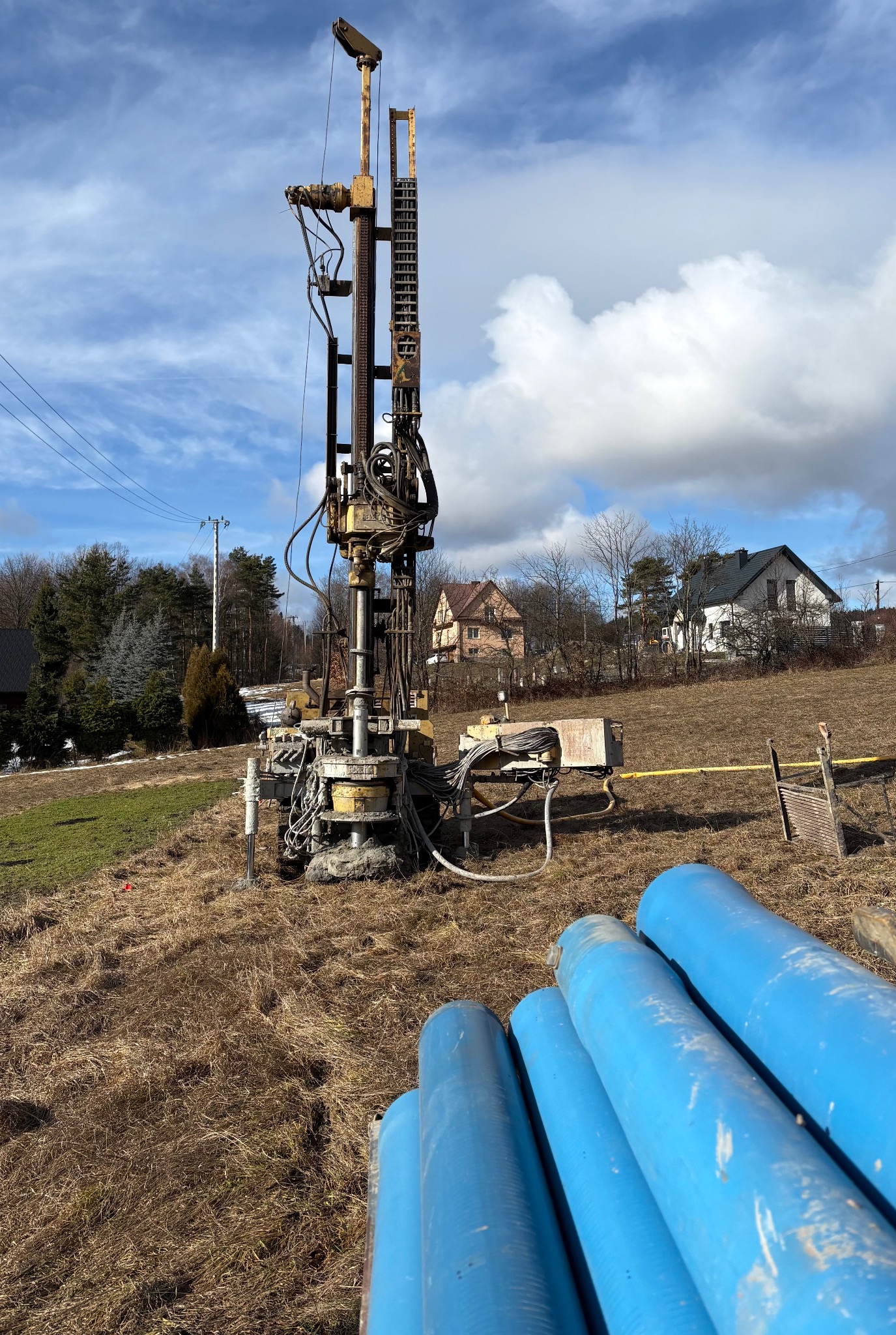 Wiertnica do studni głębinowych na zewnątrz, z niebieskimi rurami w pierwszym planie. W tle domy i błękitne niebo z chmurami. Widok z dołu.