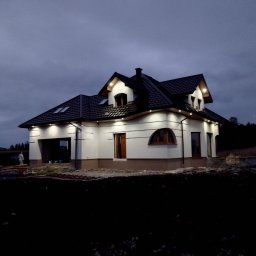 Michal Jopowicz Usługi remontowo budowlane - Nowoczesny, oświetlony dom jednorodzinny z ciemnym dachem i białymi ścianami, sfotografowany o zmierzchu. Widoczne elementy wykończeniowe i teren wokół domu w trakcie prac.
