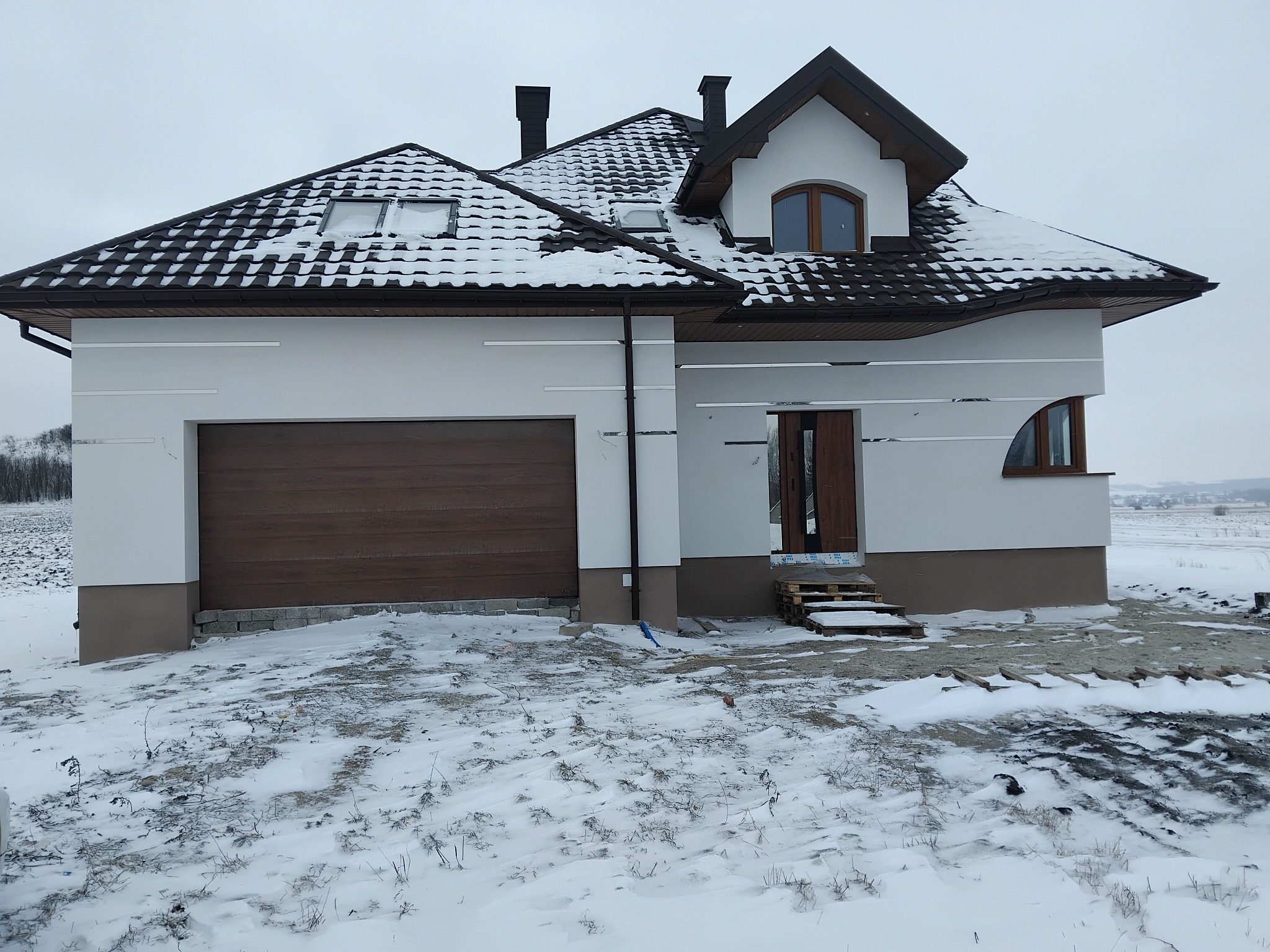 Nowoczesny dom jednorodzinny z brązową bramą garażową i dachem pokrytym śniegiem, w zimowej scenerii. Widoczne detale elewacji i otoczenie budynku.
