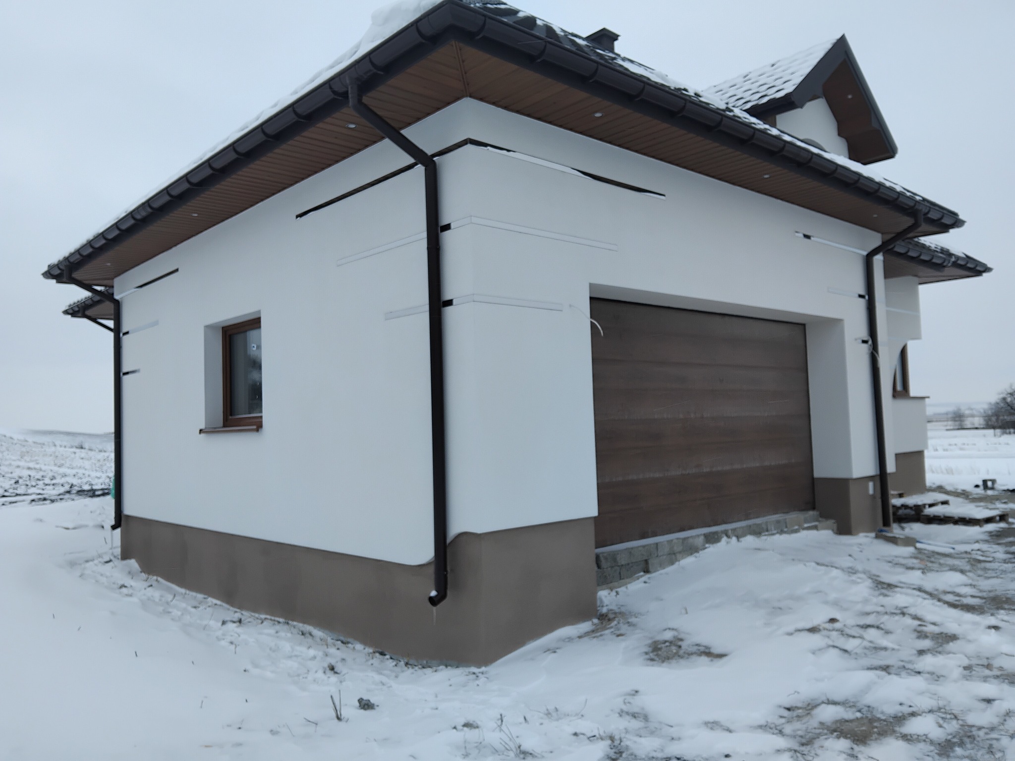 Biały dom z brązową bramą garażową i ciemnym dachem w zimowej scenerii. Widoczne rynny i fragment okna. Śnieg pokrywa ziemię.