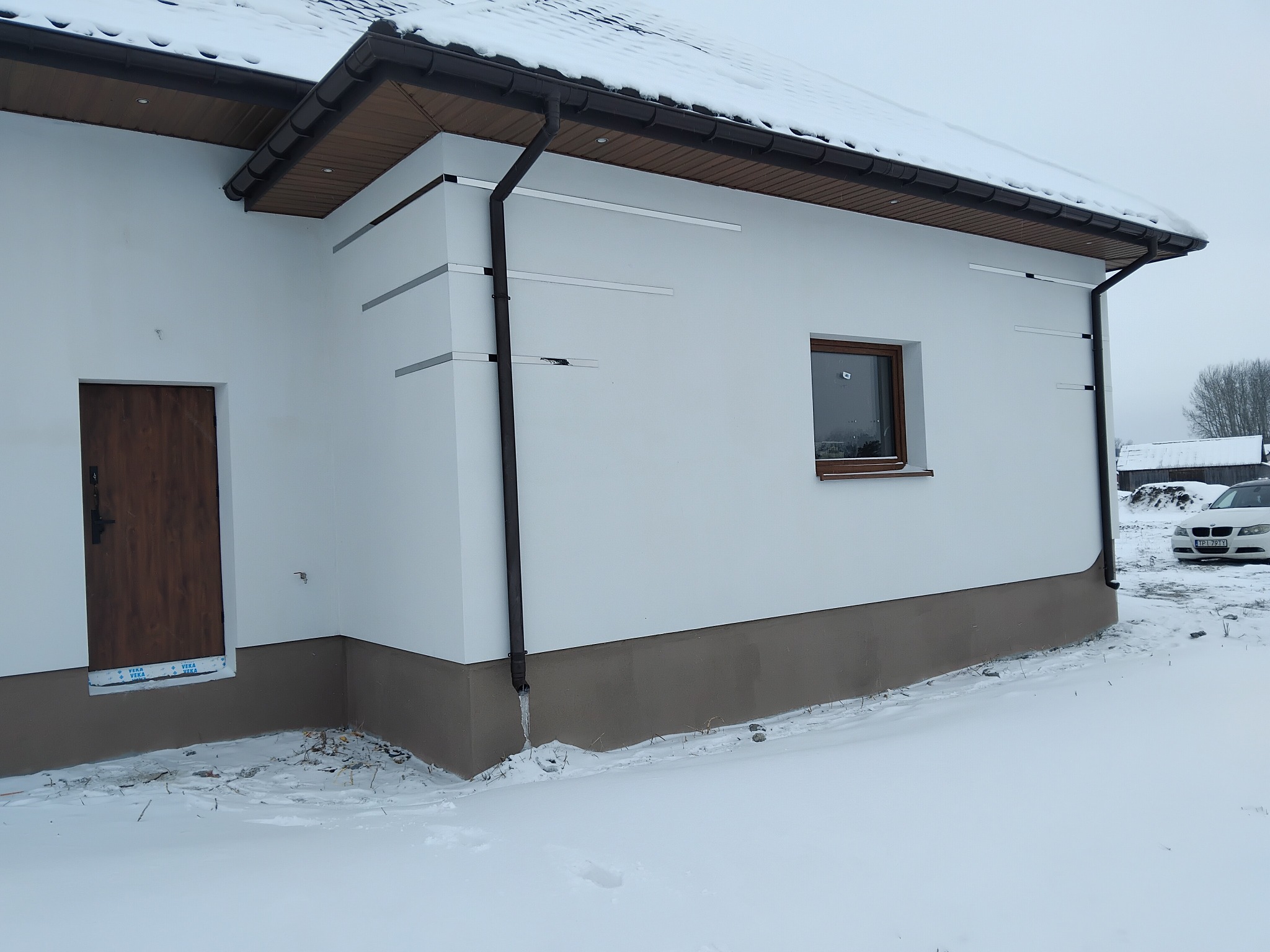 Biała elewacja domu z brązowymi akcentami (okno, drzwi, rynny) w zimowej scenerii. Widoczne listwy montażowe i fragment ośnieżonego dachu. Częściowo ośnieżony teren.