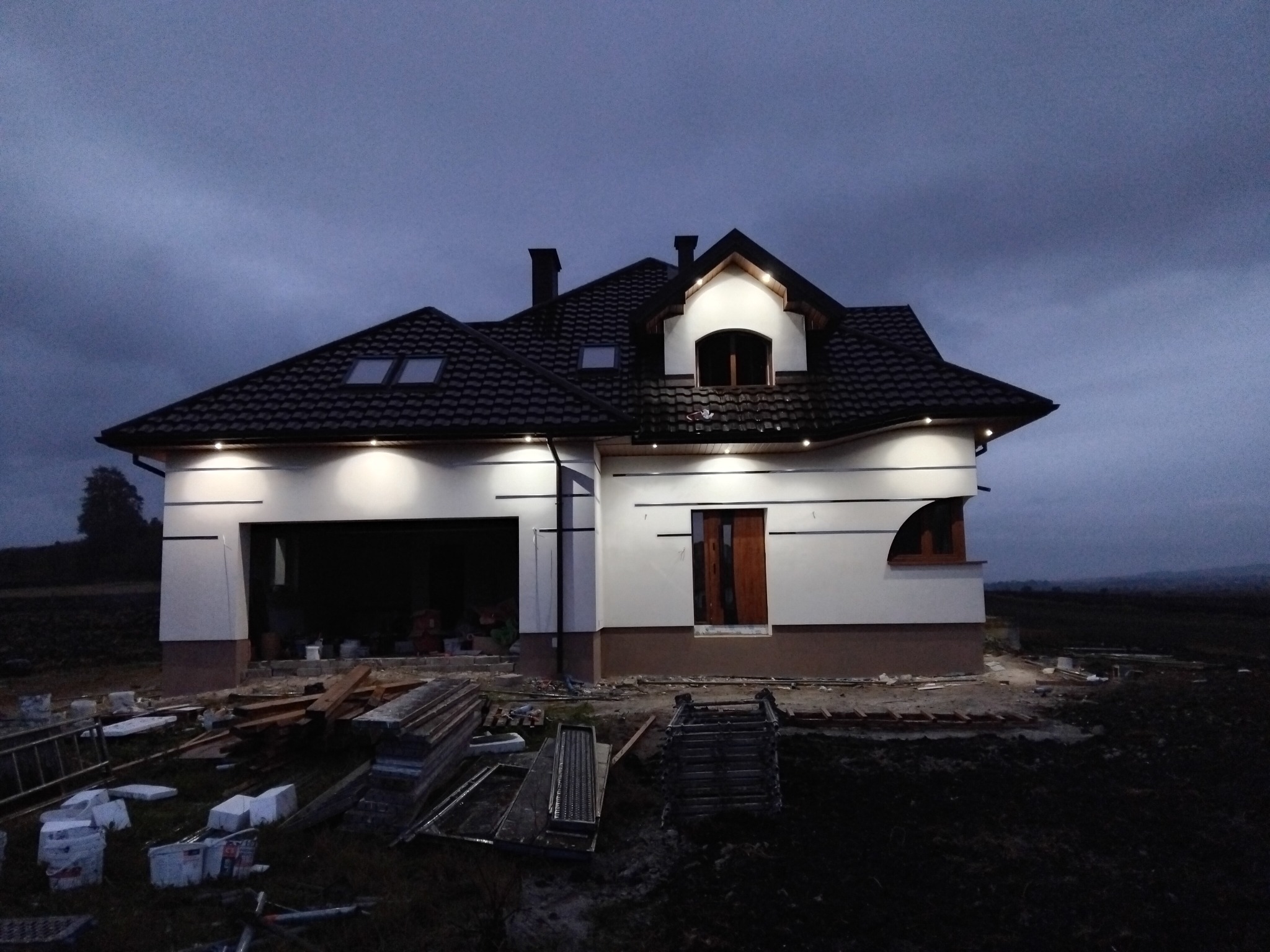 Nowoczesny dom jednorodzinny z ciemnym dachem i białymi ścianami, oświetlony punktowo, w trakcie prac wykończeniowych, widoczny w pochmurny dzień.