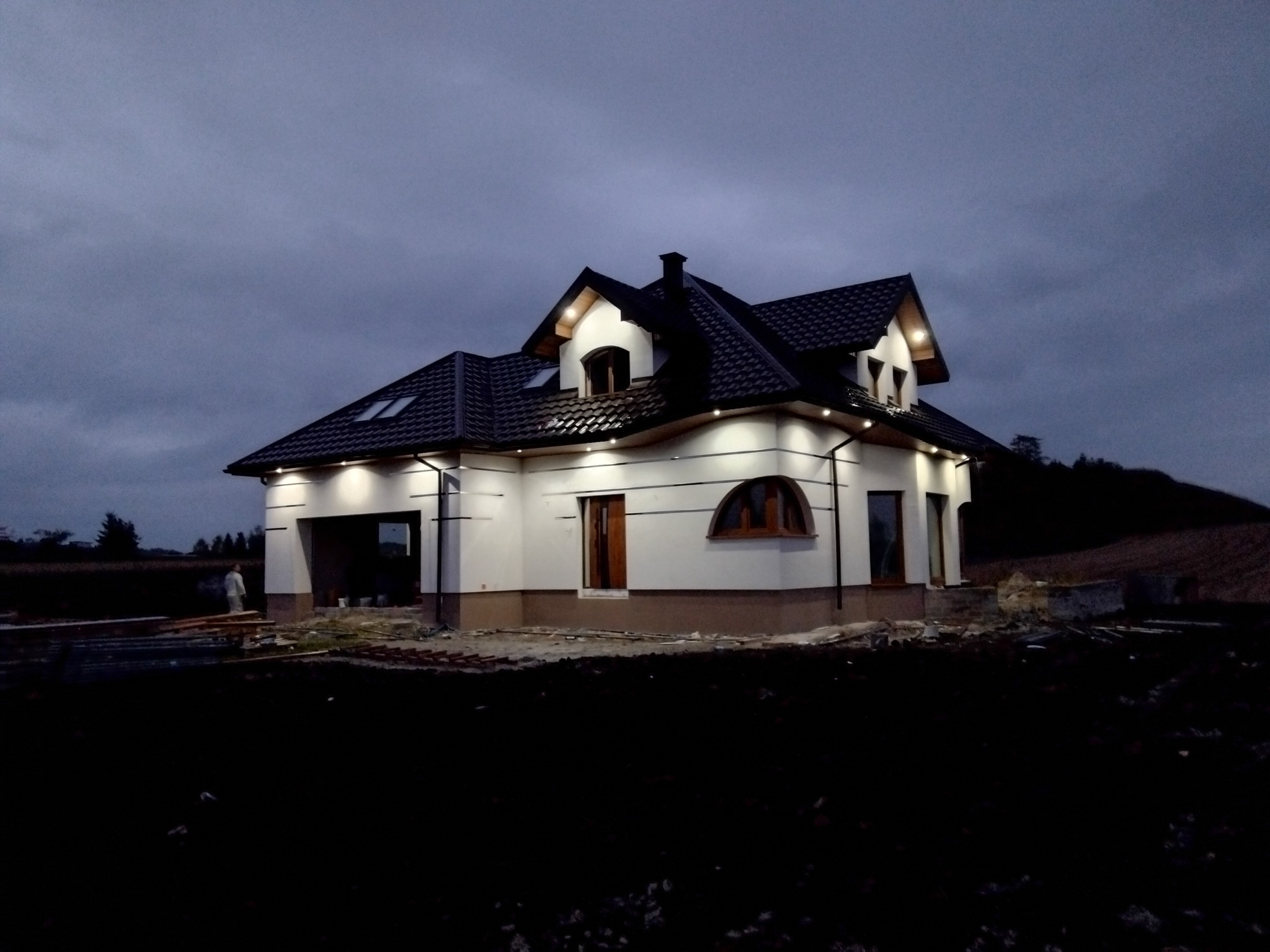 Nowoczesny, oświetlony dom jednorodzinny z ciemnym dachem i białymi ścianami, sfotografowany o zmierzchu. Widoczne elementy wykończeniowe i teren wokół domu w trakcie prac.