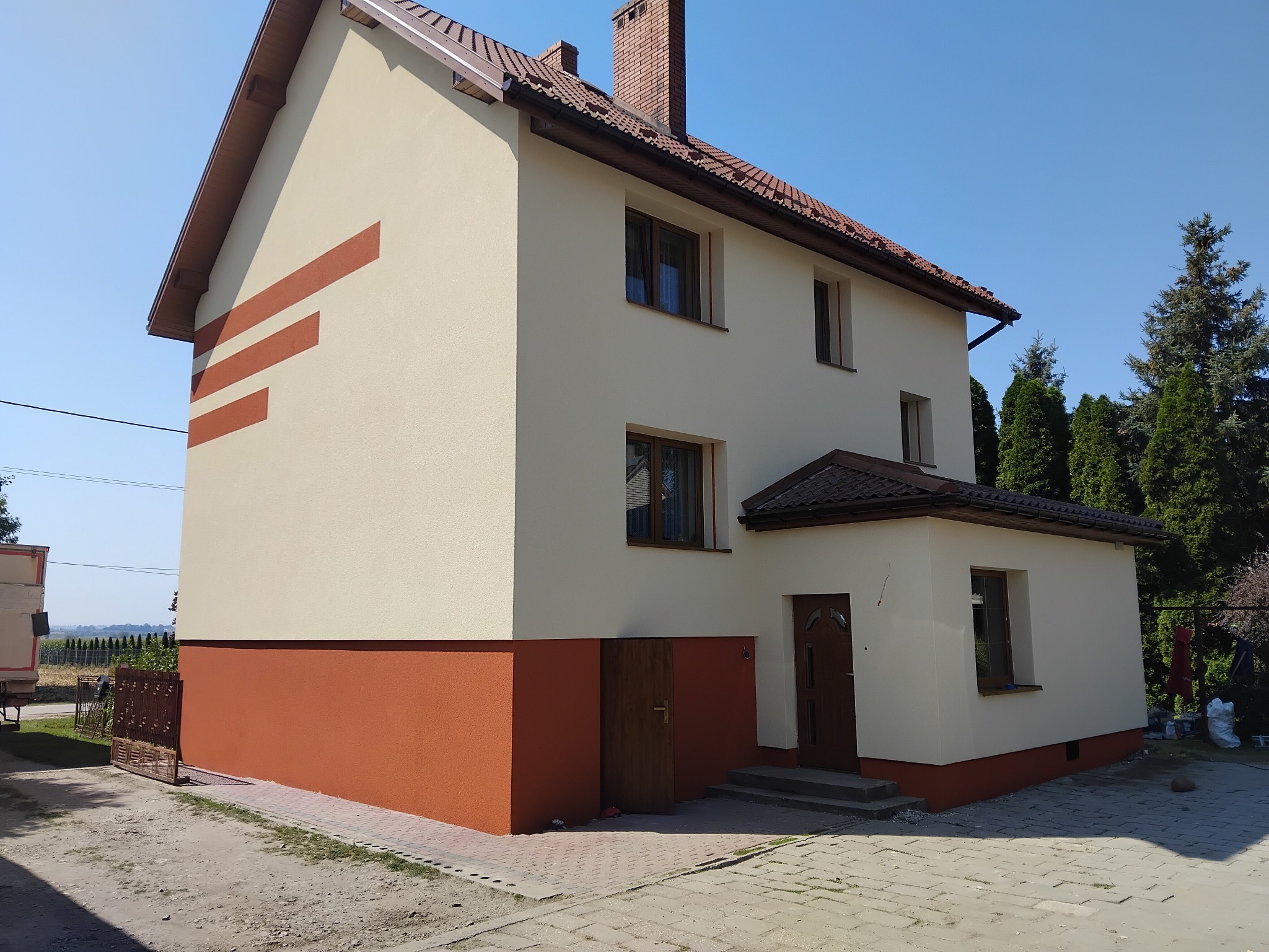 Dwukondygnacyjny dom z beżową elewacją i brązowym cokołem, ozdobiony poziomymi pasami. Widoczny komin z cegły i brązowa dachówka. Ujęcie w słoneczny dzień.