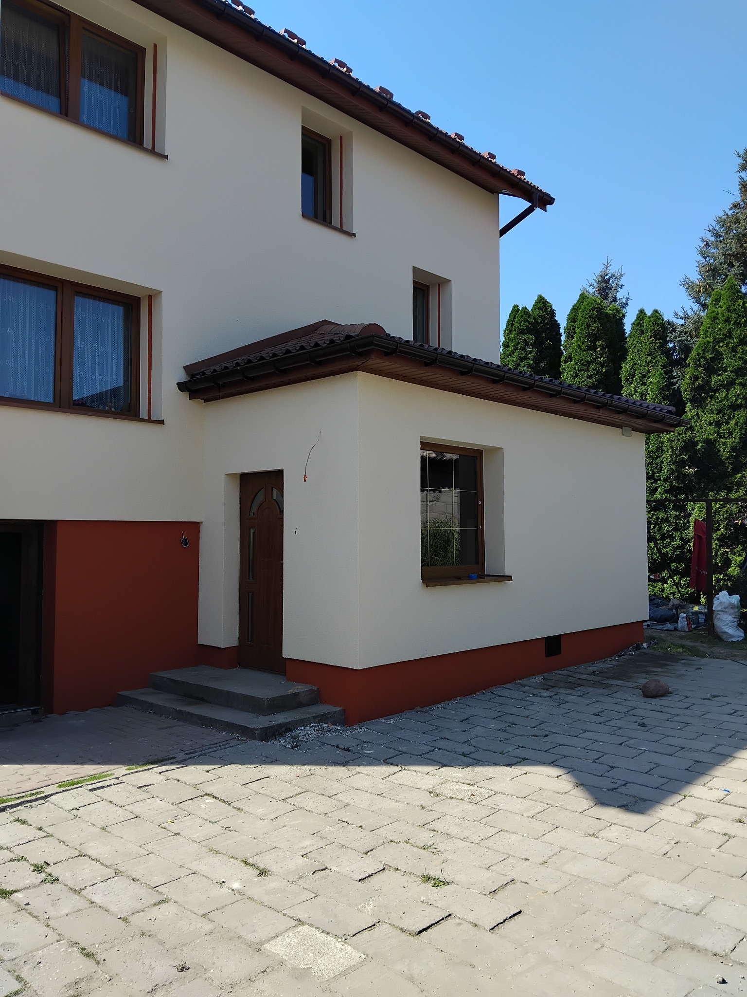 Dwukondygnacyjny dom z jasną elewacją i brązowymi akcentami, widoczny z zewnątrz, z brukowanym podjazdem i zielonymi drzewami w tle, w słoneczny dzień.