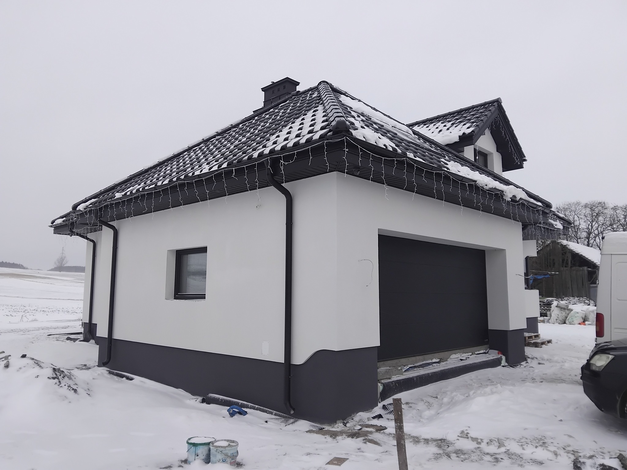 Otynkowany, parterowy budynek z garażem w zimowej scenerii. Świeżo pomalowana elewacja w odcieniach bieli i szarości, czarny dach z dekoracyjnym oświetleniem.