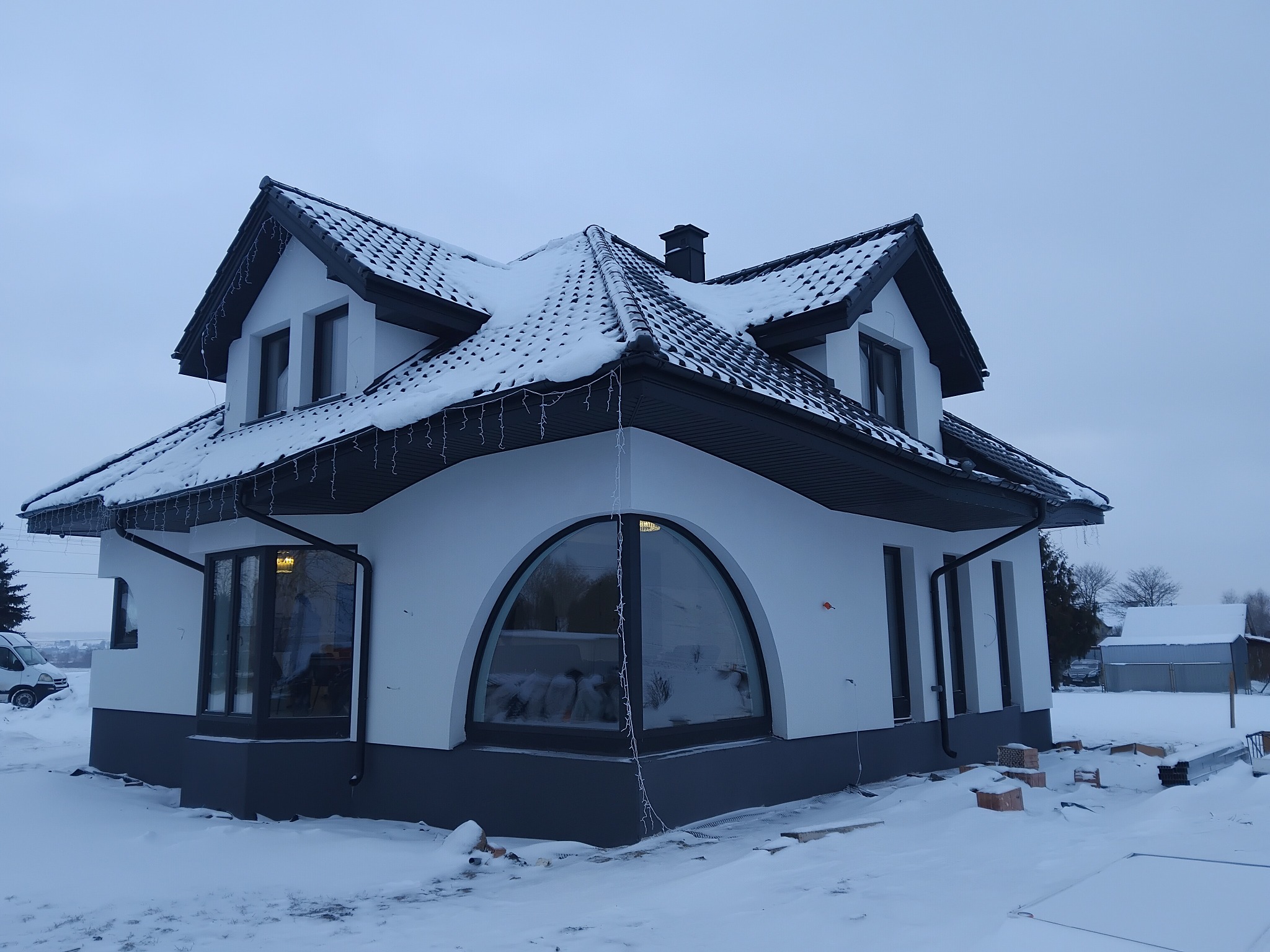Dwukondygnacyjny dom z ciemną dachówką i dużym, łukowym oknem, częściowo pokryty śniegiem. Widoczne dekoracje świąteczne i materiały budowlane na zewnątrz.