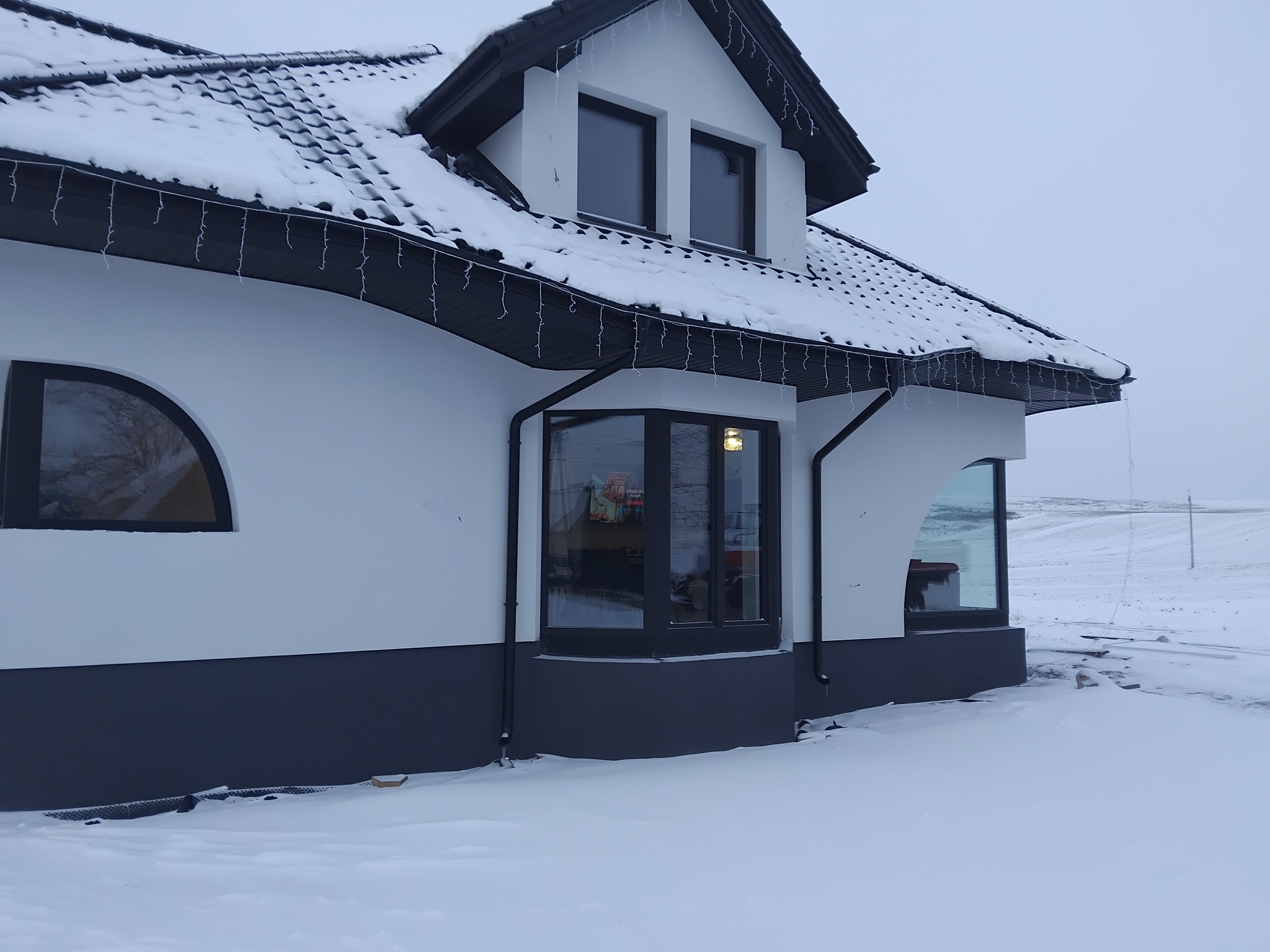 Biały dom z czarnym dachem pokrytym śniegiem, widok z boku w zimowej scenerii. Dekoracyjne lampki zwisają z dachu. Dolna część elewacji pomalowana na ciemny kolor.