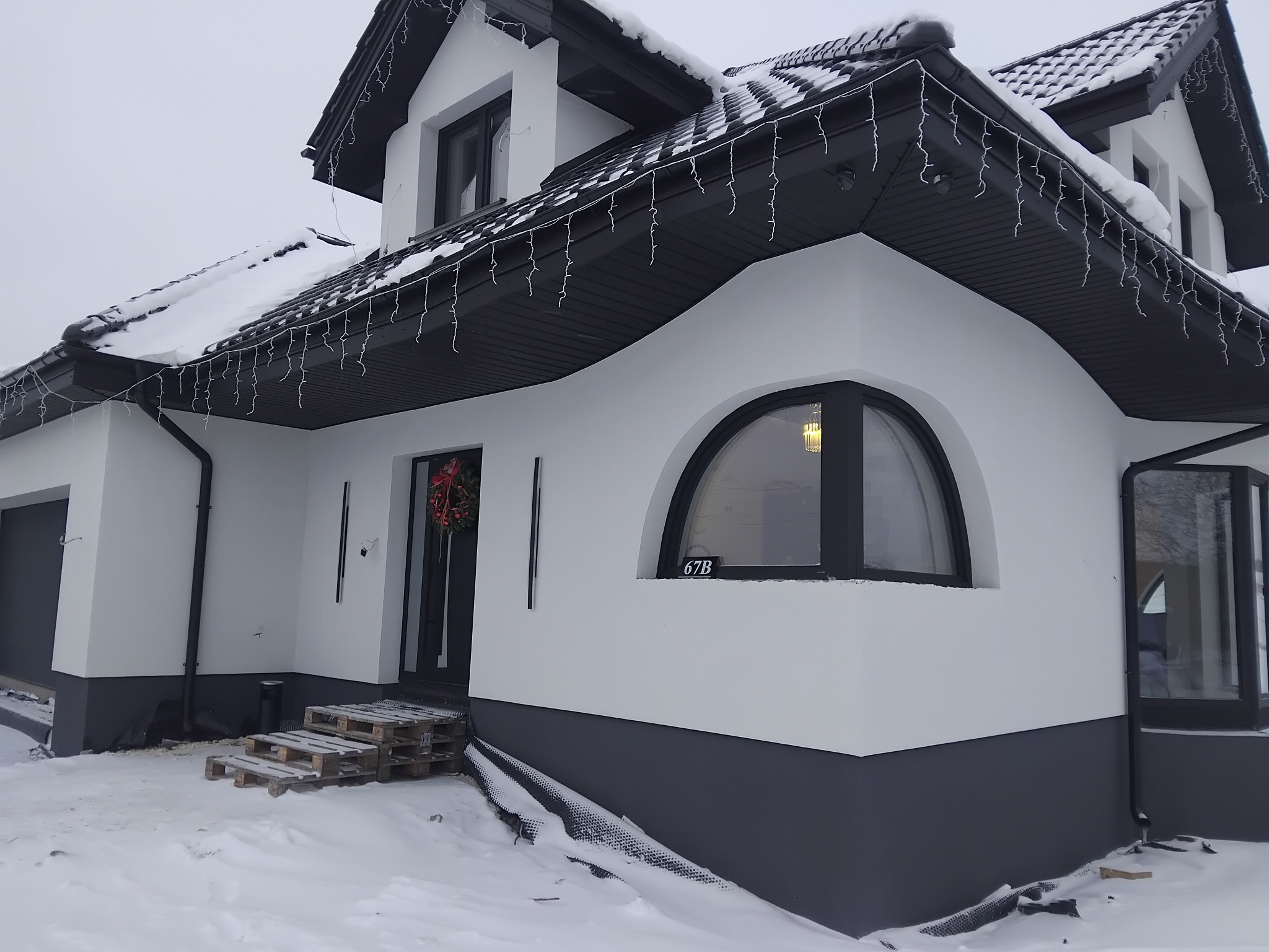 Nowoczesny dom jednorodzinny w zimowej scenerii, z białą elewacją i grafitowymi akcentami, dekorowany świątecznym wieńcem i lampkami. Widoczne palety jako tymczasowe schody.