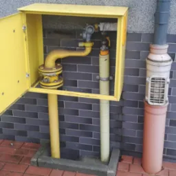 Żółta skrzynka gazowa z otwartymi drzwiczkami, widoczna instalacja gazowa wewnątrz, na tle ściany z szarej cegły i czerwonej kostki brukowej w Kleosinie.