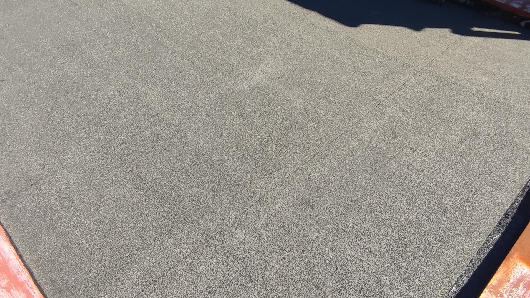 Szary, płaski dach pokryty papą, widoczny z góry, z fragmentem cienia i krawędzią dachu w prawym dolnym rogu. Minimalistyczny kadr.