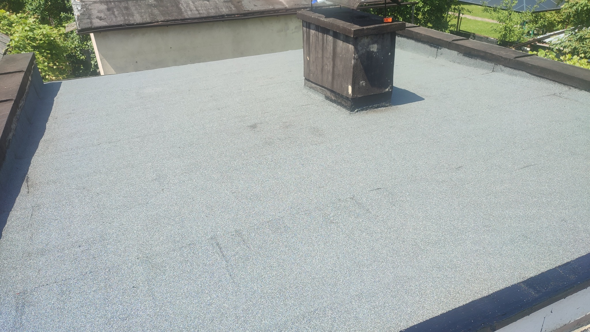 Szary, płaski dach pokryty papą z widocznym kominem i obróbkami blacharskimi. Widok z góry, w tle zieleń drzew.