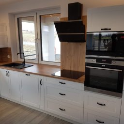 Zakład stolarski Paweł Śmigielski - Nowoczesna kuchnia na wymiar z białymi frontami, drewnianym blatem i czarnymi akcentami, w tym okap i zlew. Widoczne okno z widokiem na zewnątrz.