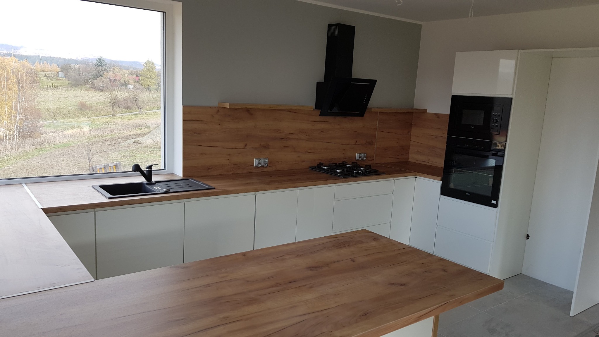 Nowoczesna kuchnia z białymi szafkami i drewnianym blatem, okapem i piekarnikiem w zabudowie. Widok na krajobraz za oknem. Uwagę przykuwa drewniany blat imitujący naturalne drewno.