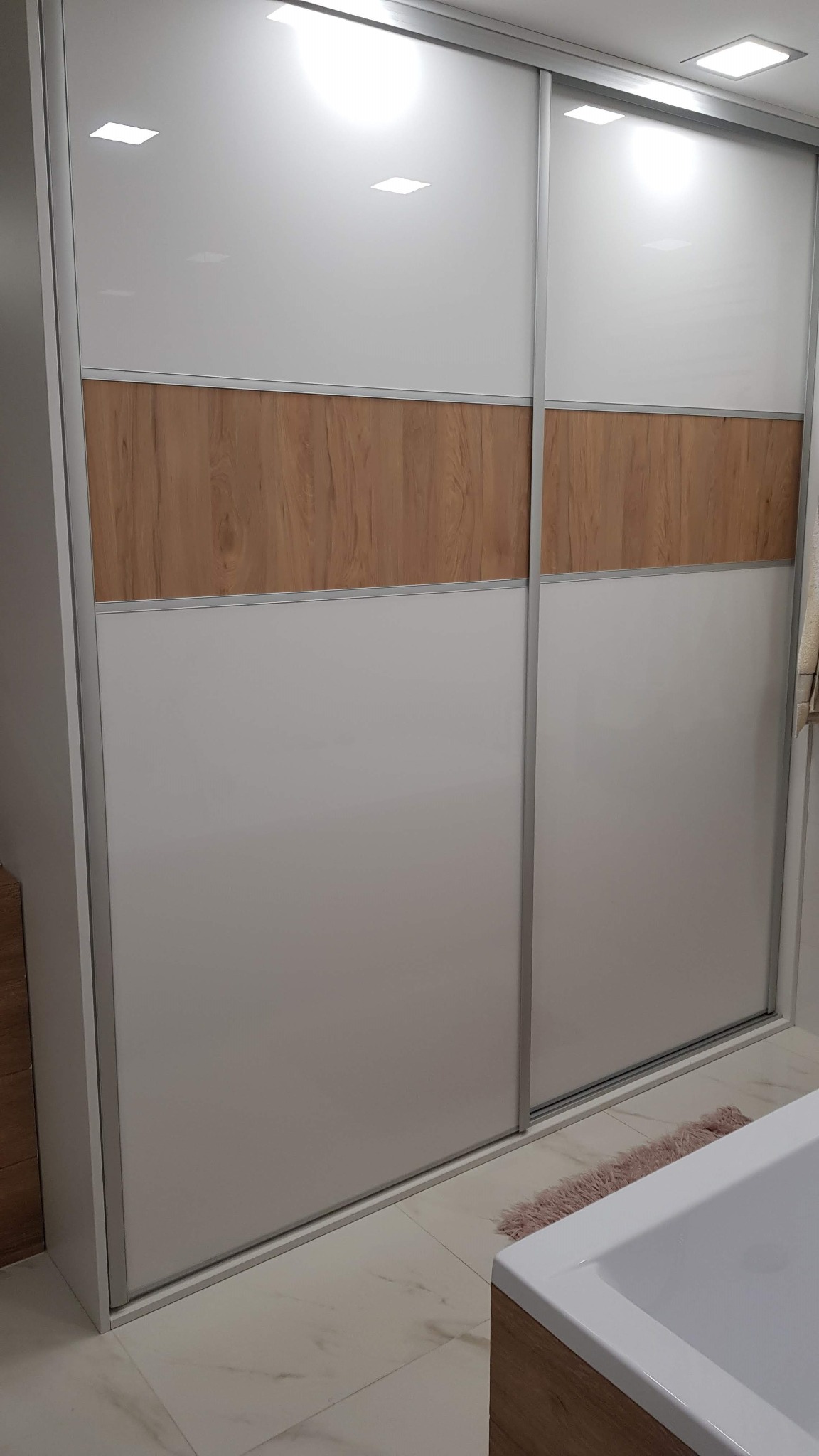 Nowoczesna szafa wnękowa z przesuwnymi drzwiami, biało-drewniana, z oświetleniem punktowym. Widoczny fragment łazienki z białą wanną i marmurową podłogą.