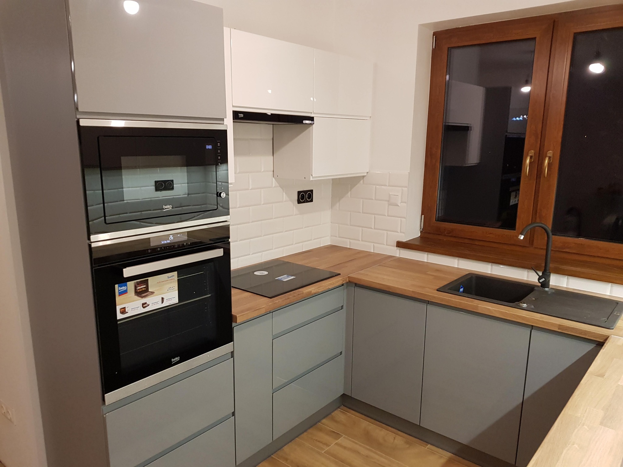 Nowoczesna kuchnia z szarymi szafkami, drewnianym blatem i białymi płytkami. Widoczny piekarnik Beko i okno z drewnianą ramą. Minimalistyczny design.