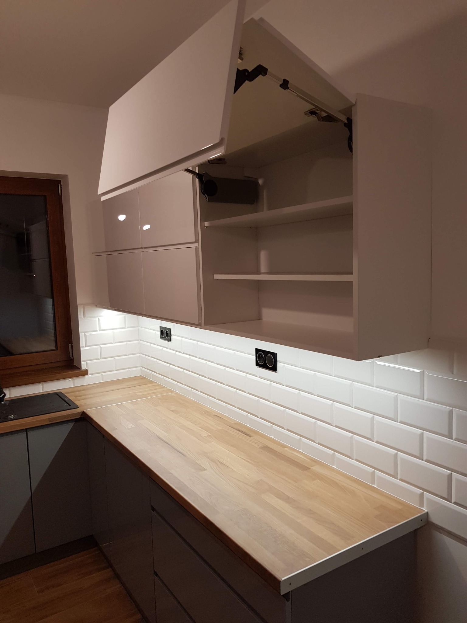 Nowoczesna kuchnia z drewnianym blatem i białymi, błyszczącymi szafkami wiszącymi. Jedna szafka otwarta, ukazująca półki. Białe płytki na ścianie i podświetlenie LED.