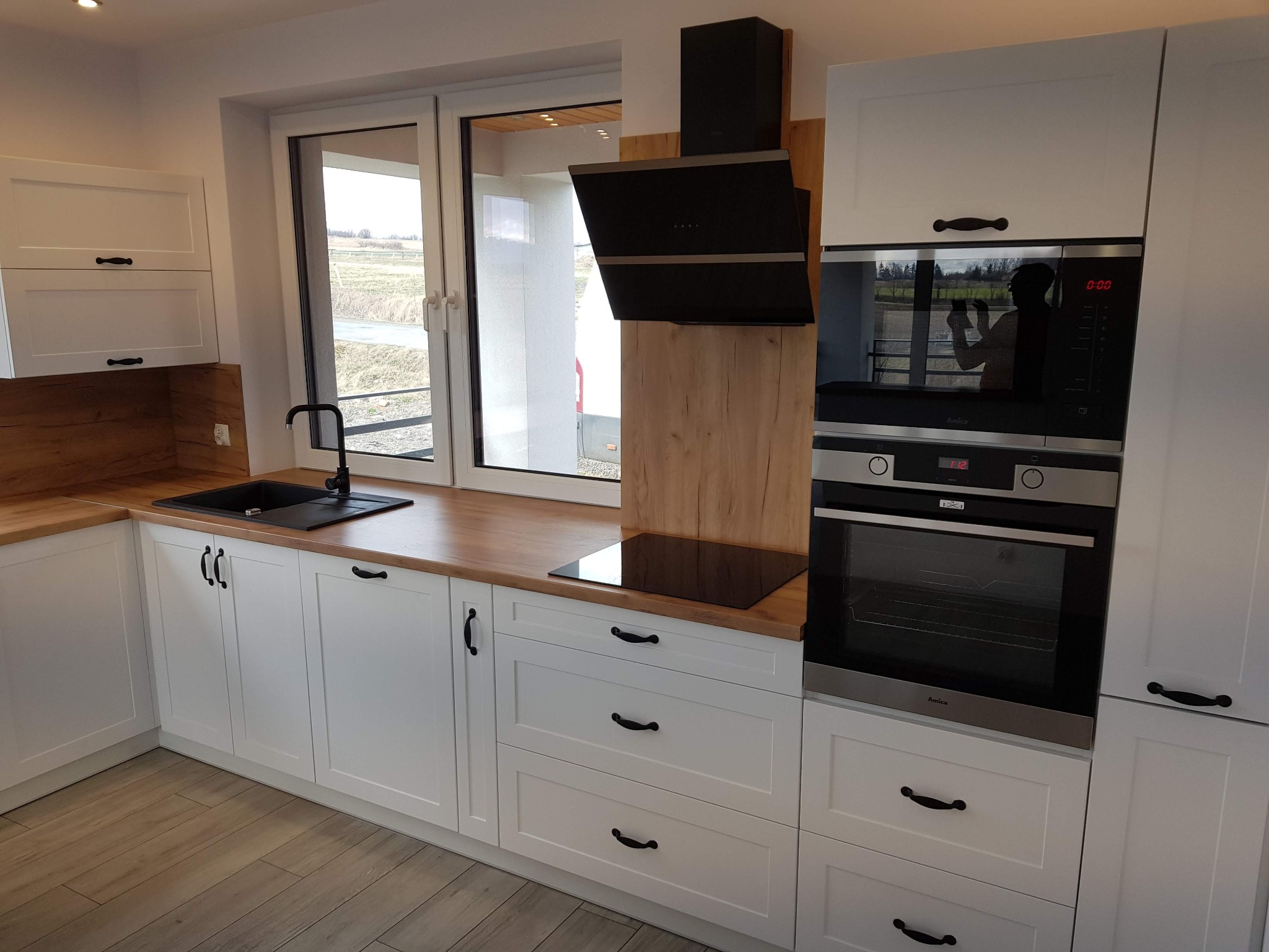 Nowoczesna kuchnia na wymiar z białymi frontami, drewnianym blatem i czarnymi akcentami, w tym okap i zlew. Widoczne okno z widokiem na zewnątrz.