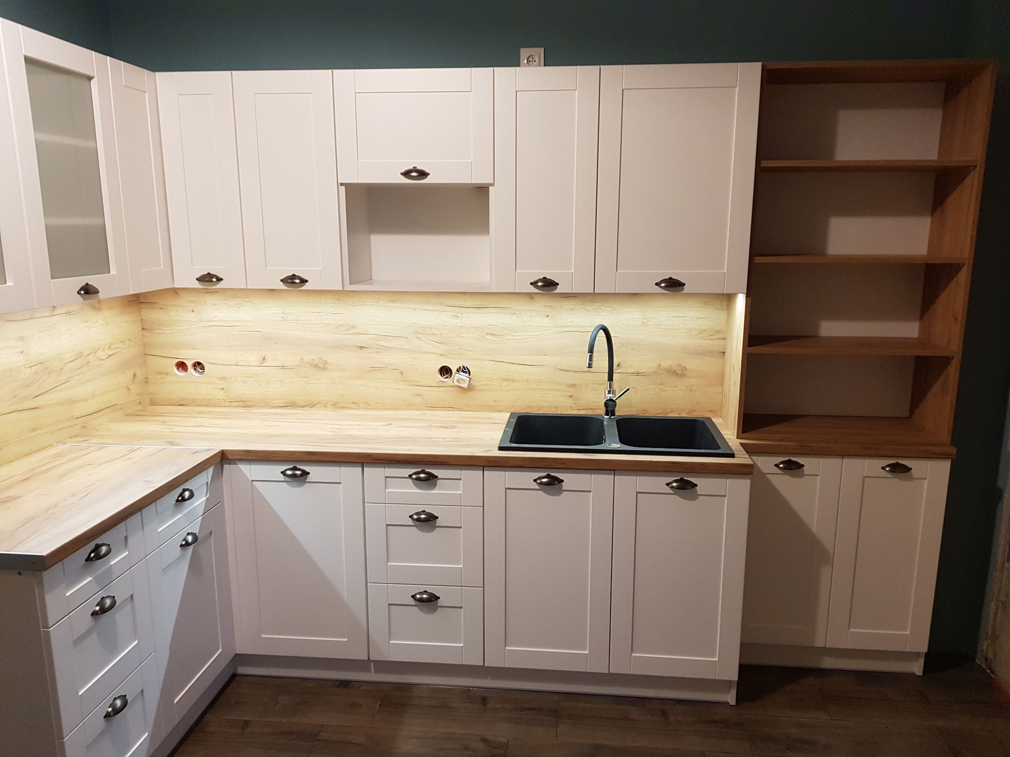 Jasna kuchnia z drewnianym blatem i czarnym zlewozmywakiem. Fronty szafek białe, frezowane, z ciemnymi uchwytami. Na ścianie drewnopodobna okładzina. Widoczna półka.