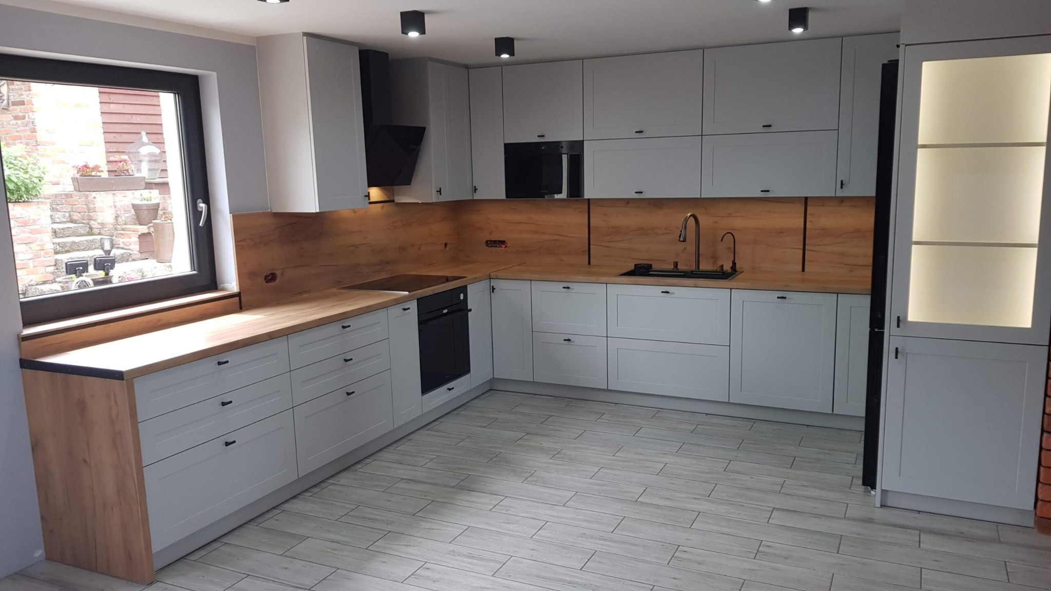 Jasnoszara kuchnia na wymiar z drewnianymi blatami i oświetleniem punktowym, zrealizowana w Radomsku. Nowoczesny design i funkcjonalne rozwiązania.