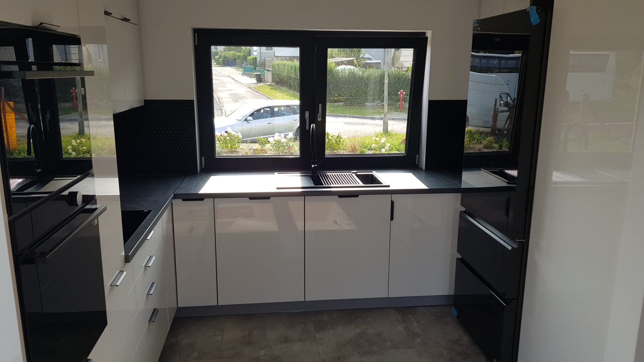 Nowoczesna kuchnia z białymi szafkami i czarnym blatem, widok na ulicę przez okno nad zlewem. Minimalistyczny design, błyszczące fronty, czarne akcenty.