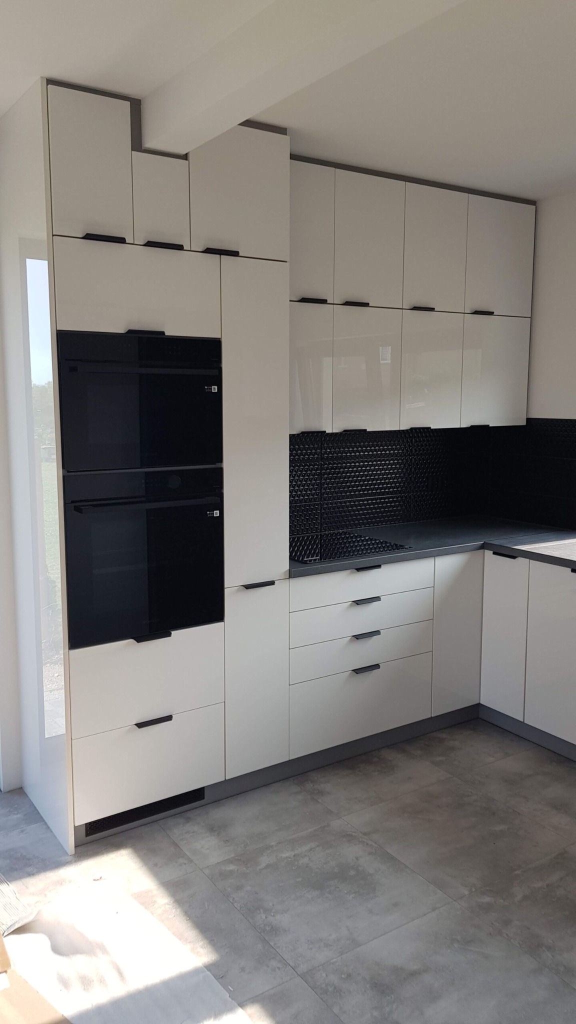 Biała, nowoczesna kuchnia z czarnymi akcentami: piekarnik, płyta i uchwyty. Błyszczące fronty szafek górnych i dolnych, szara podłoga. Minimalistyczny design.