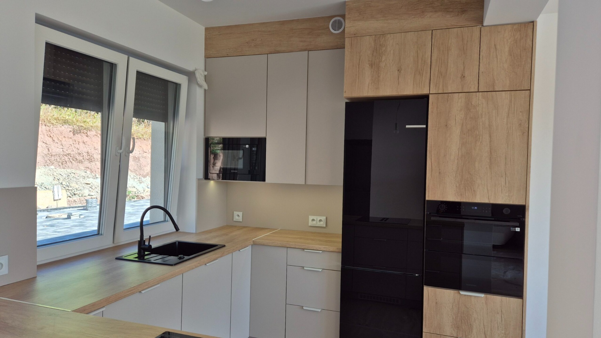 Nowoczesna kuchnia z szarymi szafkami i drewnianym blatem, czarny zlew pod oknem, zabudowana lodówka i piekarnik. Minimalistyczny design z oświetleniem LED.