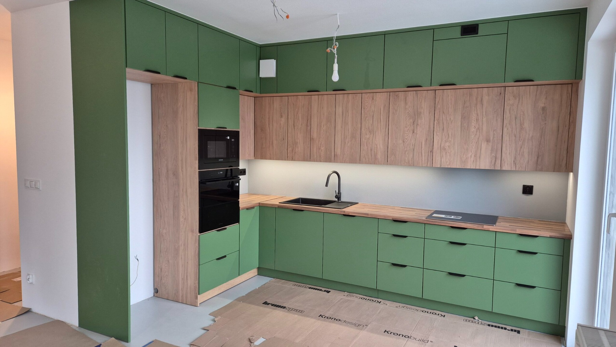 Nowoczesna kuchnia z zielonymi szafkami i drewnianym blatem. Uwagę zwraca minimalistyczny design i oświetlenie LED pod szafkami wiszącymi. Na podłodze karton ochronny.
