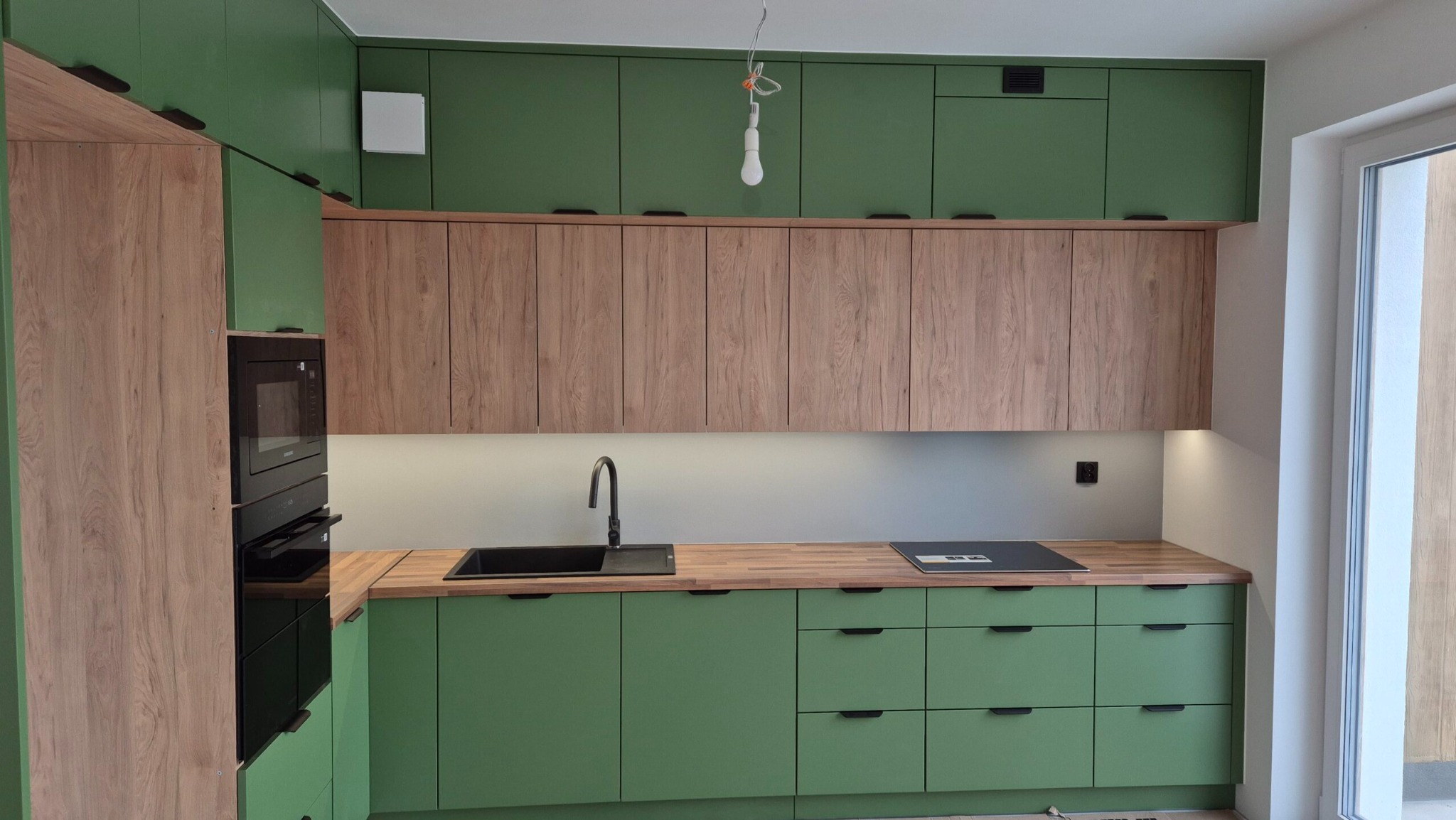 Nowoczesna kuchnia na wymiar z zielonymi szafkami dolnymi i drewnopodobnymi górnymi. Czarny zlew i płyta indukcyjna na drewnianym blacie. Wisząca żarówka bez klosza.