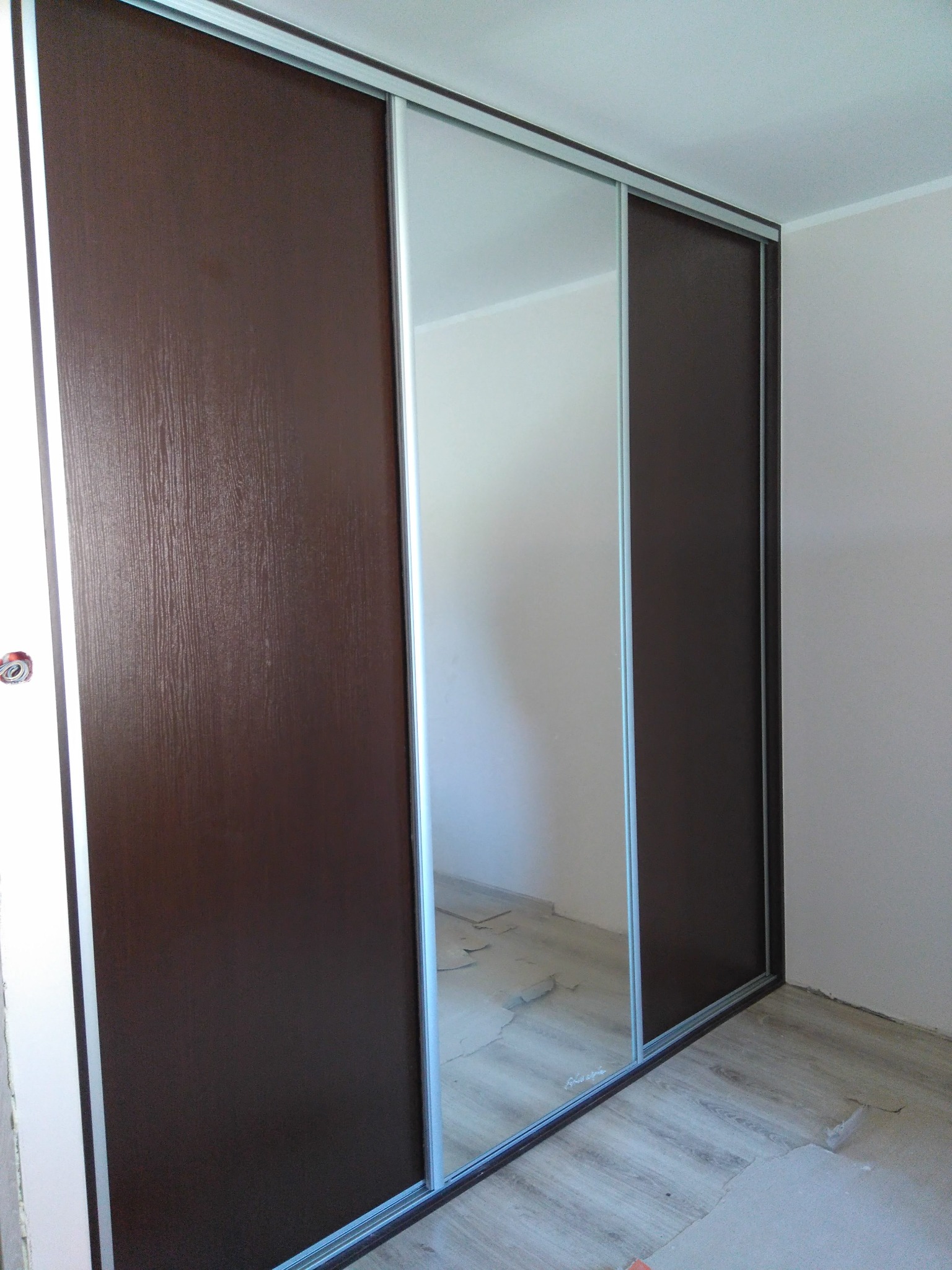 Szafa wnękowa z przesuwnymi drzwiami: brązowe panele z teksturą drewna i lustrzane środkowe drzwi. Widoczne srebrne ramy i fragment podłogi w trakcie wykończenia.
