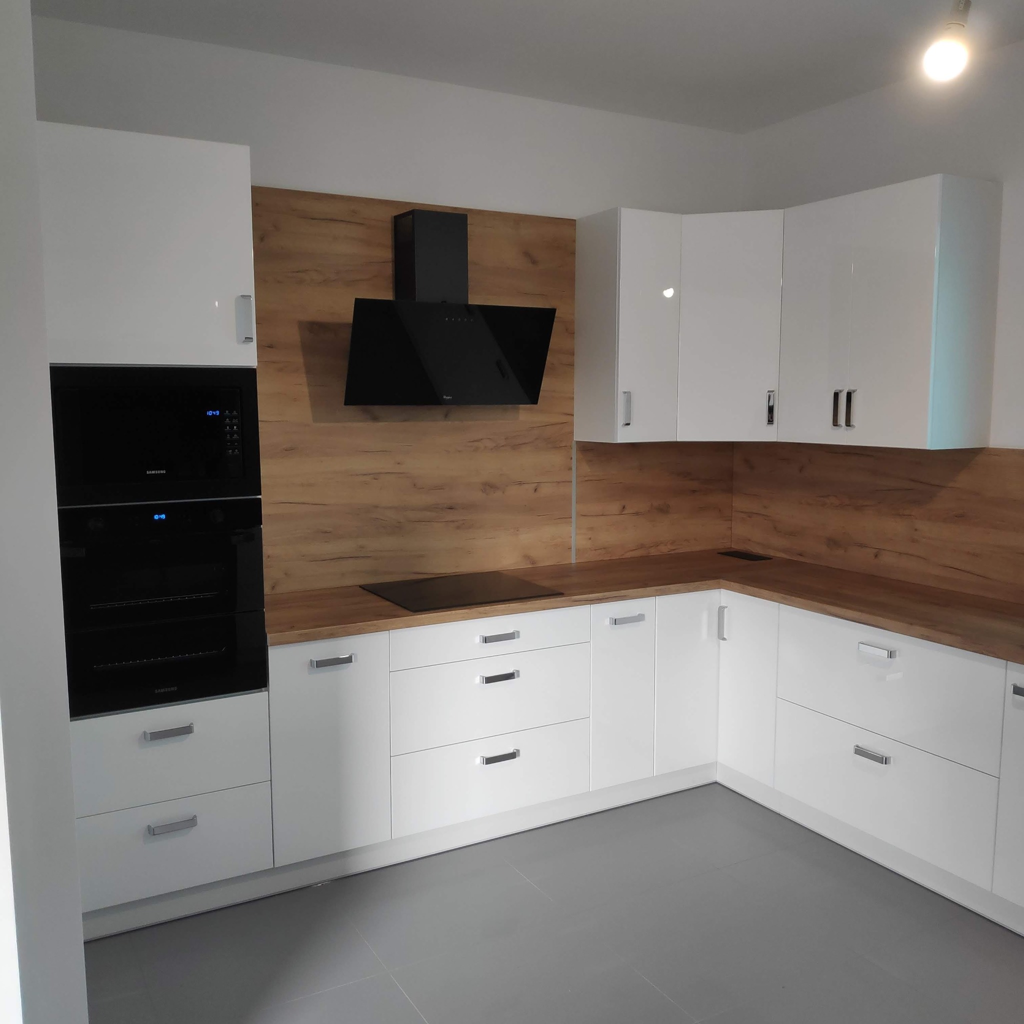Nowoczesna kuchnia z białymi, lakierowanymi frontami, drewnianym blatem i panelem ściennym. Czarny okap i zabudowany sprzęt AGD. Minimalistyczne uchwyty.