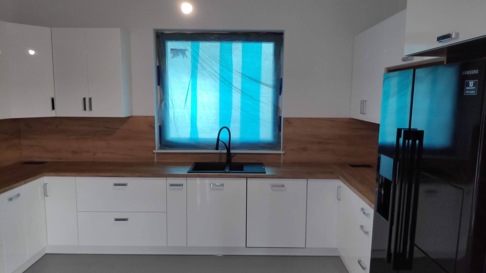 Nowoczesna kuchnia z białymi, lakierowanymi frontami, drewnianym blatem i czarnym zlewem. Okno zabezpieczone folią malarską. Widoczna lodówka Samsung.