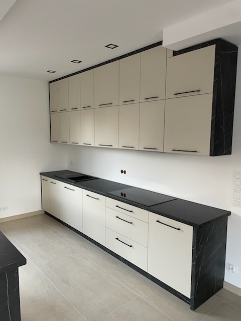 Jasna kuchnia z czarnym blatem imitującym marmur i górnymi szafkami pod sufit. Minimalistyczne uchwyty, płyta indukcyjna. Nowoczesny design.