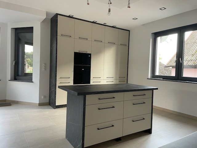 Jasna kuchnia z wyspą i zabudową po sufit, wykończona czarnym marmurem. Minimalistyczne uchwyty, duże okna wpuszczają naturalne światło. Nowoczesny design.