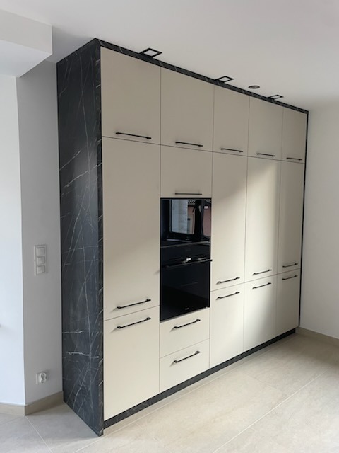 Beżowa zabudowa kuchenna z czarnymi uchwytami i piekarnikiem, wykończona marmurowym dekorem na boku, oświetlona punktowo, w nowoczesnym wnętrzu.