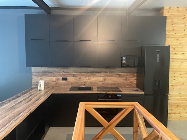 Nowoczesna kuchnia z czarnymi szafkami i drewnianym blatem. Widoczna zabudowa kuchenna, płyta indukcyjna, piekarnik i lodówka. Ściana wykończona drewnianą boazerią.