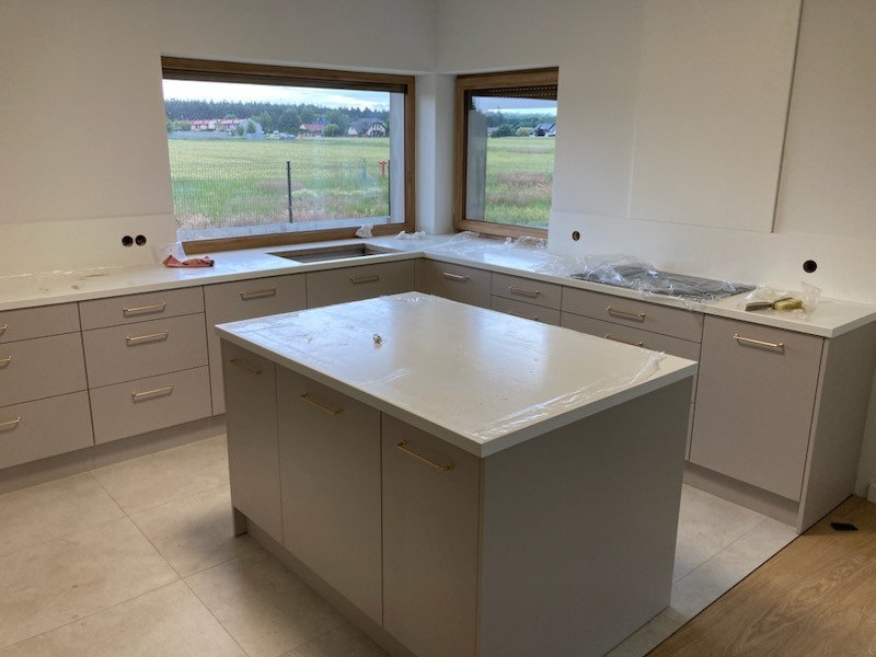 Jasna kuchnia z wyspą i widokiem na zielone pola przez okno. Minimalistyczny design, beżowe szafki z złotymi uchwytami, białe blaty. Część blatu zabezpieczona folią.