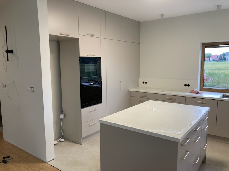 Jasna kuchnia z wyspą, zabudową pod sufit i piekarnikiem w zabudowie. Blat wyspy zabezpieczony folią. Widok na okno i fragment krajobrazu za nim.